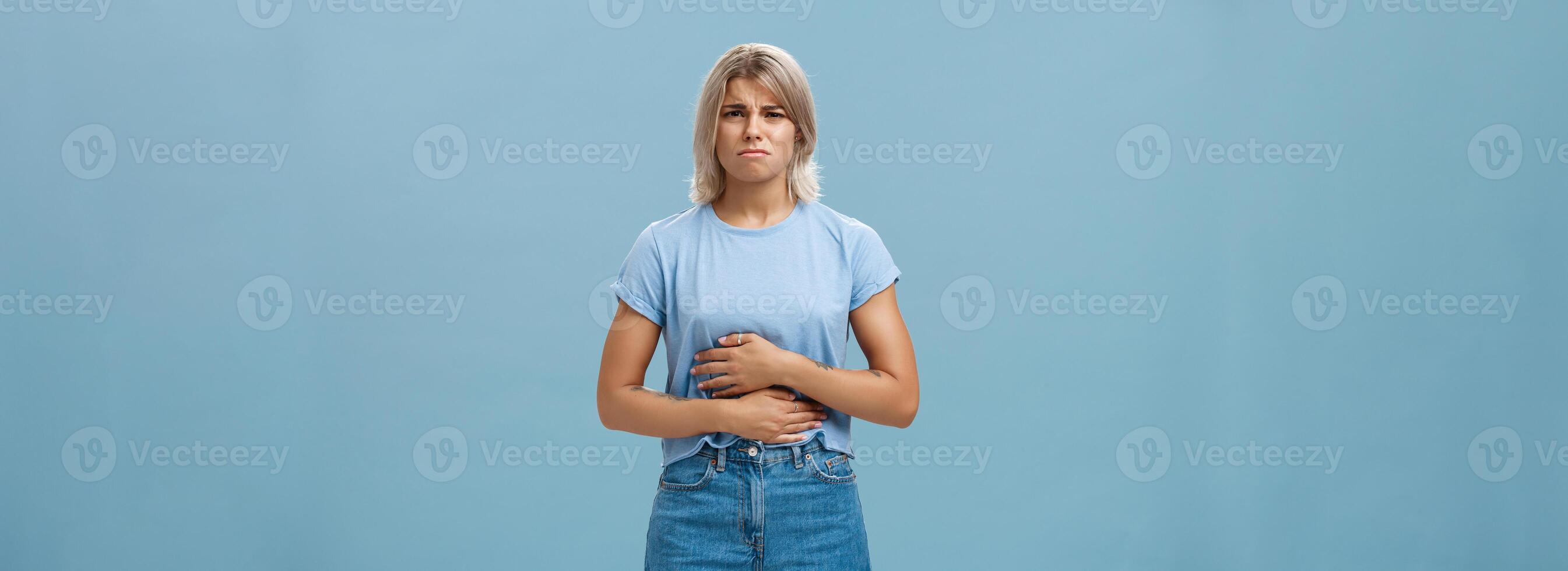 portret van niet tevreden intens aantrekkelijk jong blond meisje met tatoeages fronsen aanraken maag klagen Aan maagpijn na aan het eten rommel voedsel hebben wanorde over- blauw achtergrond foto