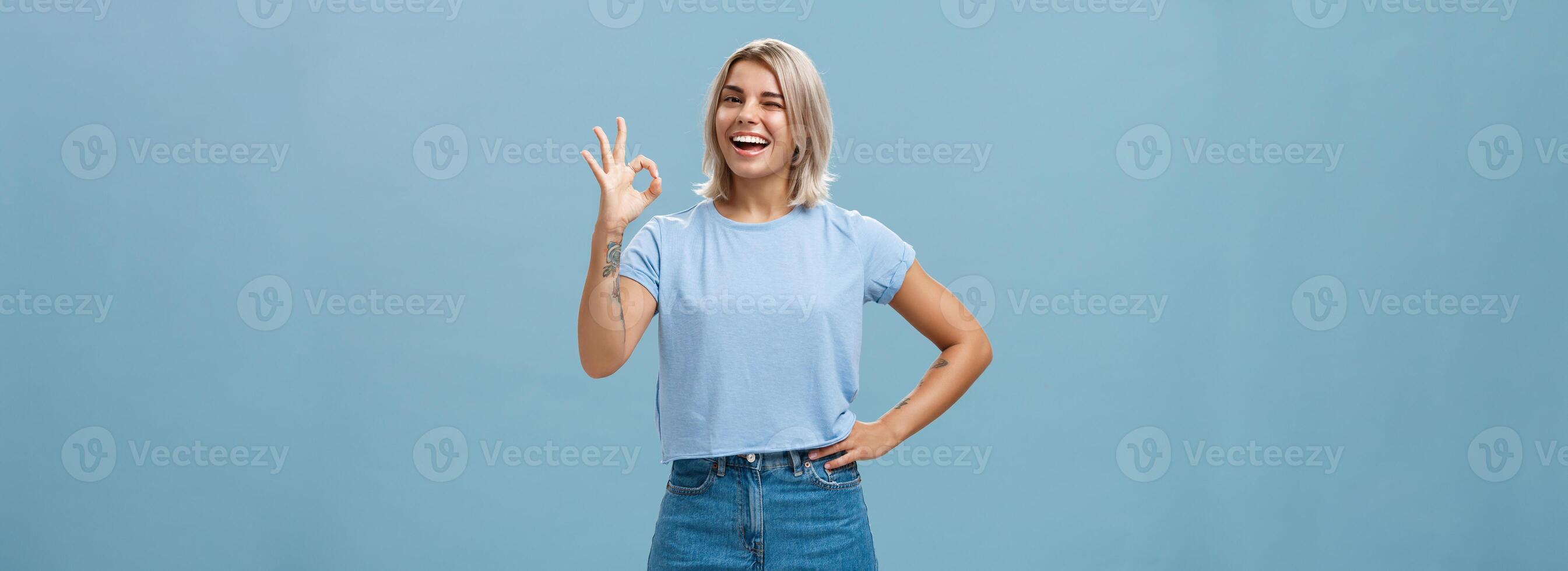 zelfverzekerd en slim meisje kreeg alles onder controle. portret van optimistisch mooi meisje met eerlijk haar- en gebruind huid knipogen tonen Oke of perfect gebaar staand over- blauw muur foto