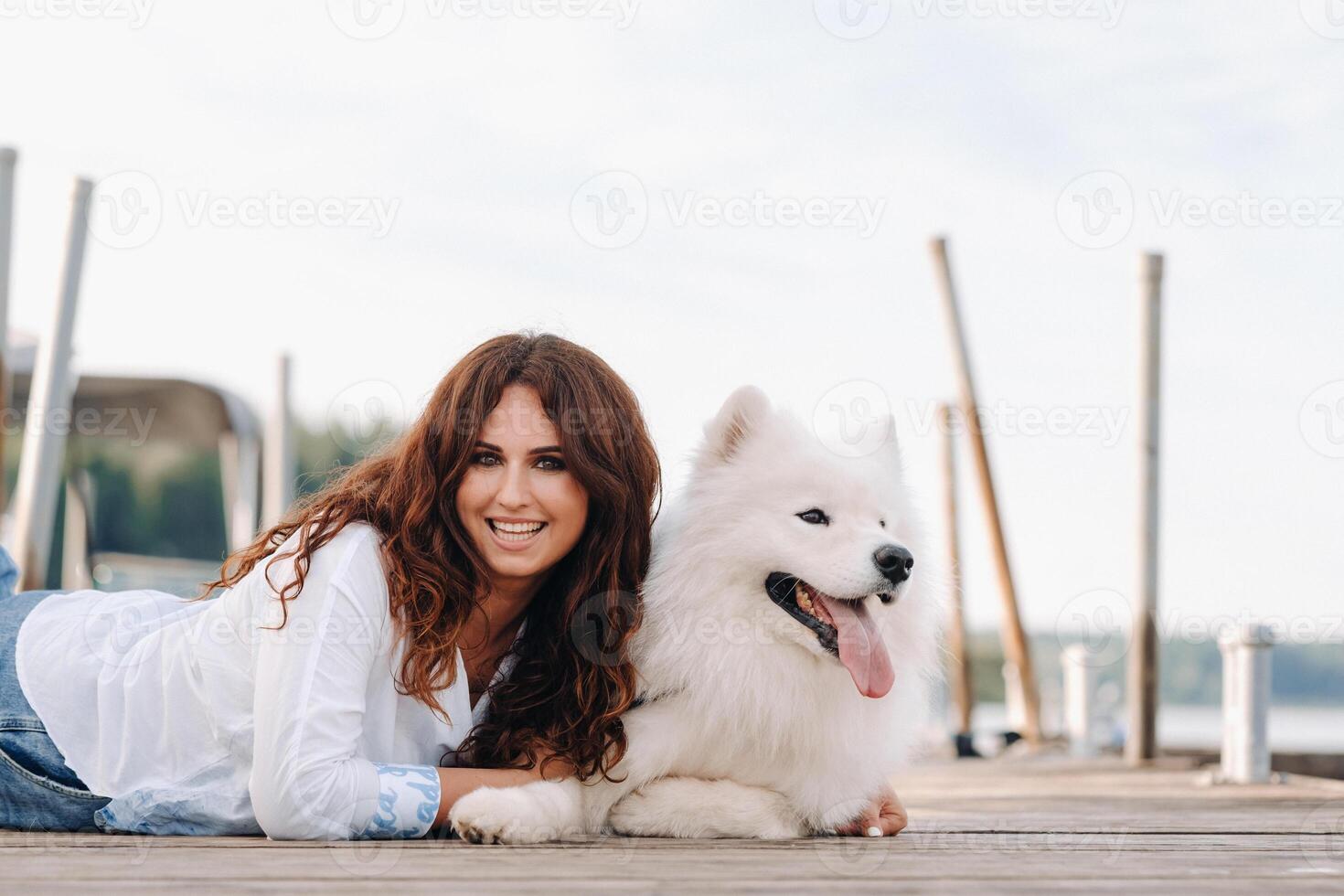 een gelukkig vrouw met een groot wit hond leugens Aan een pier in de buurt de zee Bij zonsondergang foto