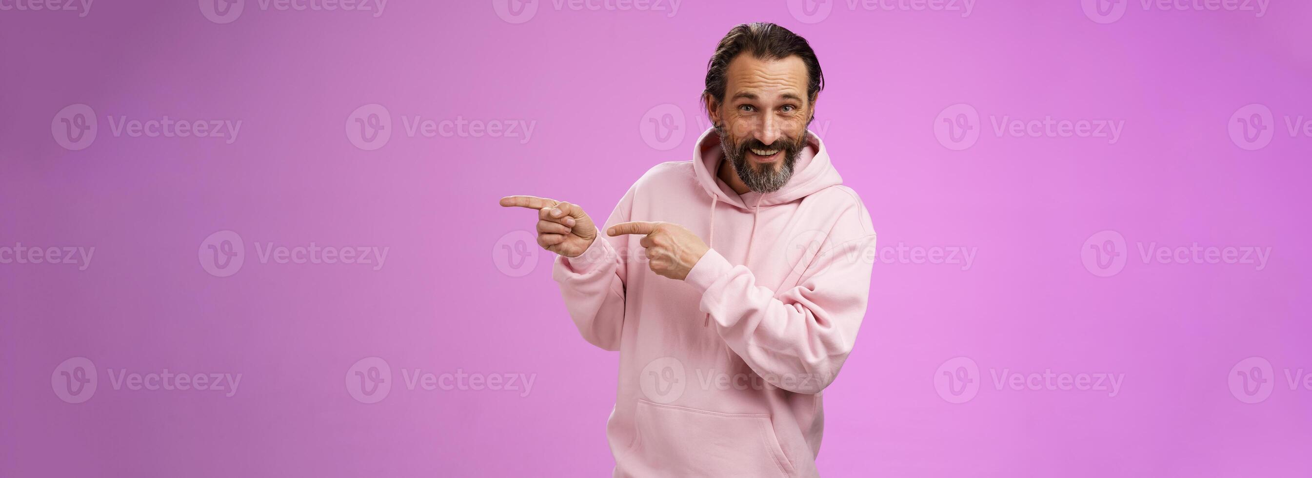 brutaal zelfverzekerd mooi macho Mens Jaren 50 in roze capuchon glimlachen breed uitnodigend toetreden richten Rechtsaf tonen interessant grappig plaats hangen uit gastvrij nemen Look, staand Purper achtergrond foto