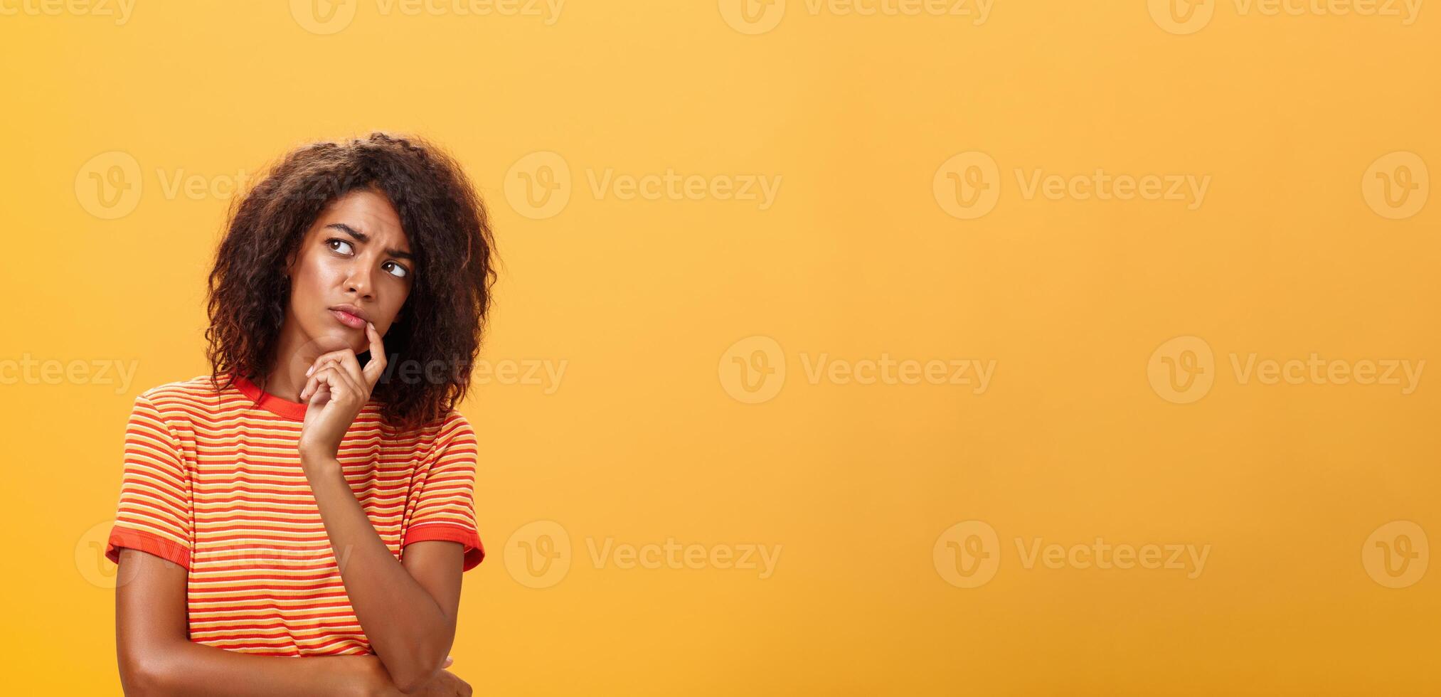 vrouw overwegen naar nemen een deel in evenement. slim gefocust en creatief mooi Afrikaanse meisje met gekruld haar- Holding vinger Aan lip staren bezorgd links, denken maken besluit over- oranje muur foto