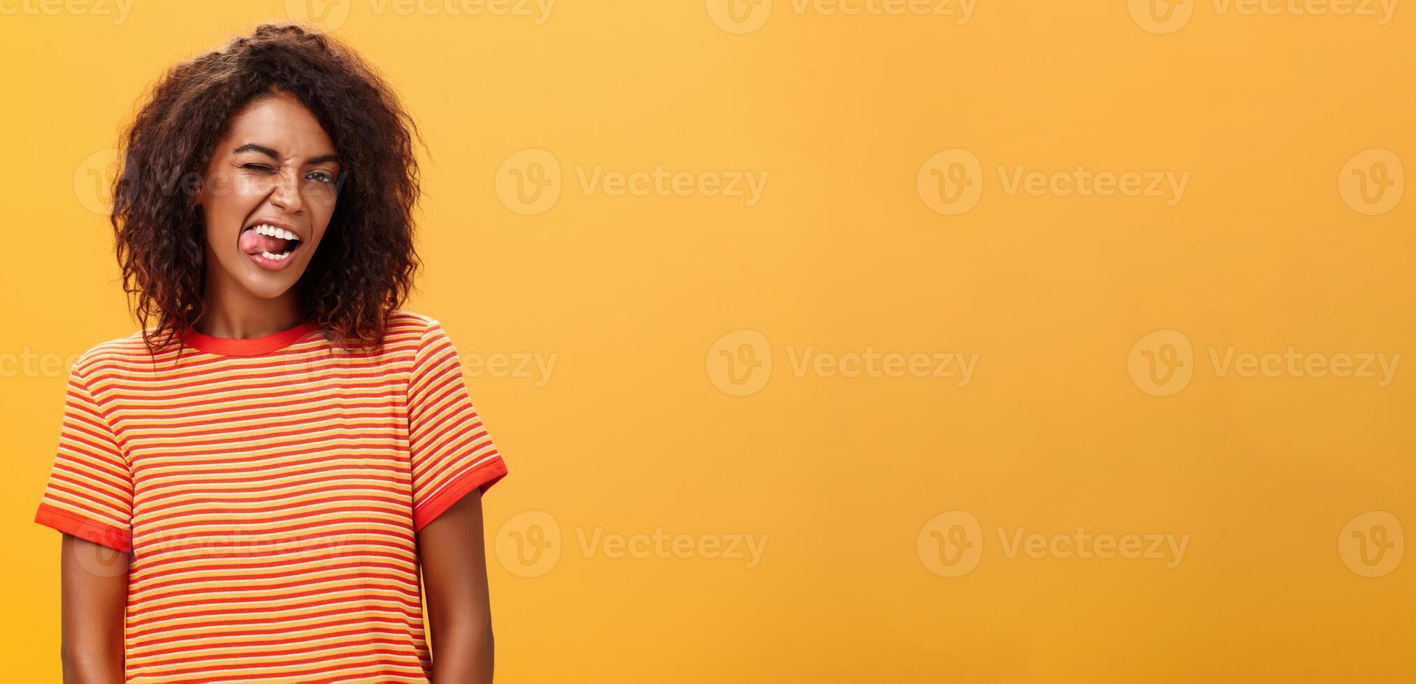 portret van gedurfd en emotioneel zelfverzekerd flirterig vrouw met afro kapsel knipogen blij tonen tong poseren zorgeloos en enthousiast tegen oranje achtergrond flirten met heet vent foto