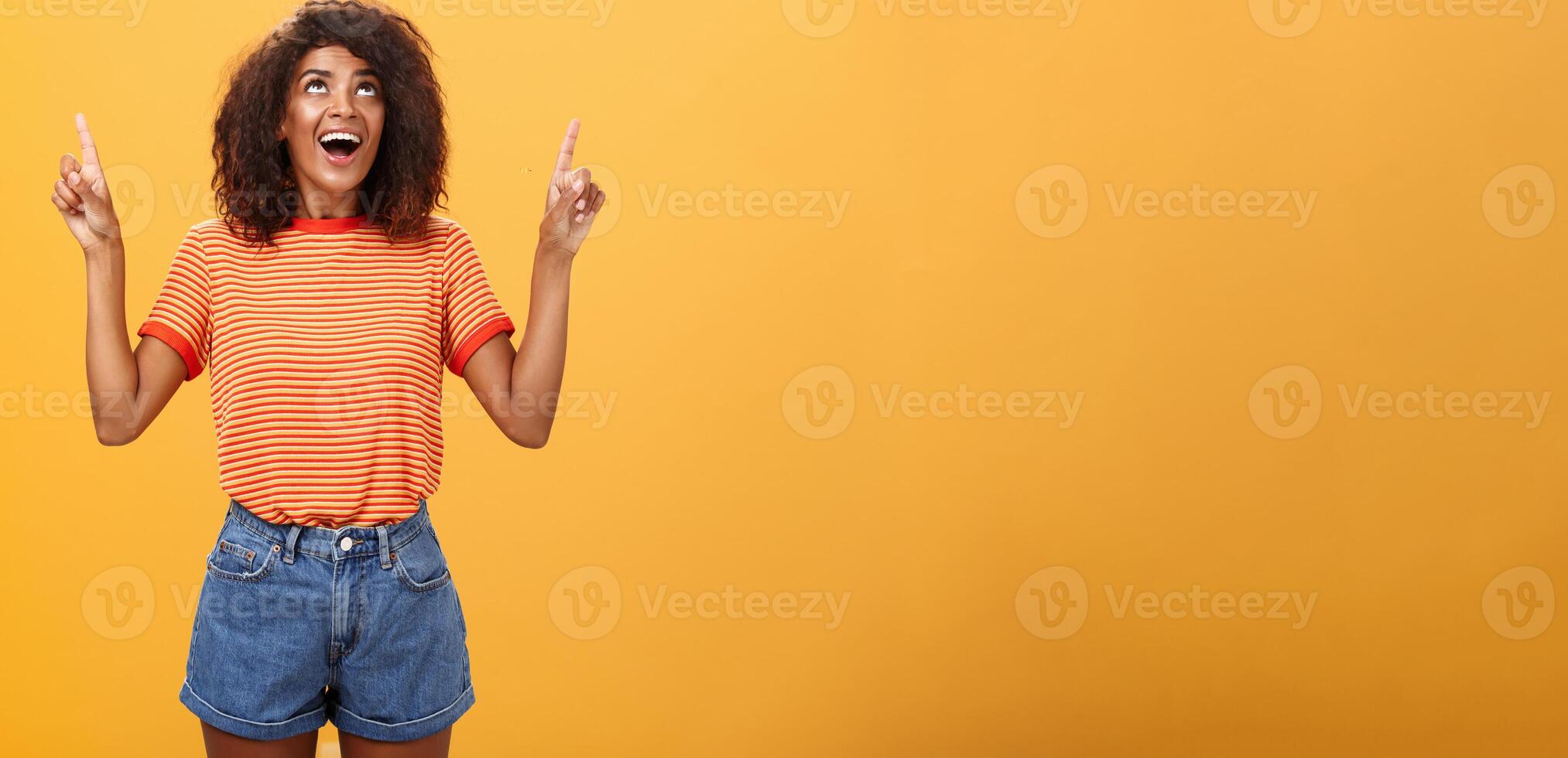 gefascineerd onder de indruk en geamuseerd mooi charmant Afro-Amerikaans vrouw met afro kapsel in modieus t-shirt en denim shorts richten en op zoek omhoog met geïnteresseerd kijken over- oranje muur foto