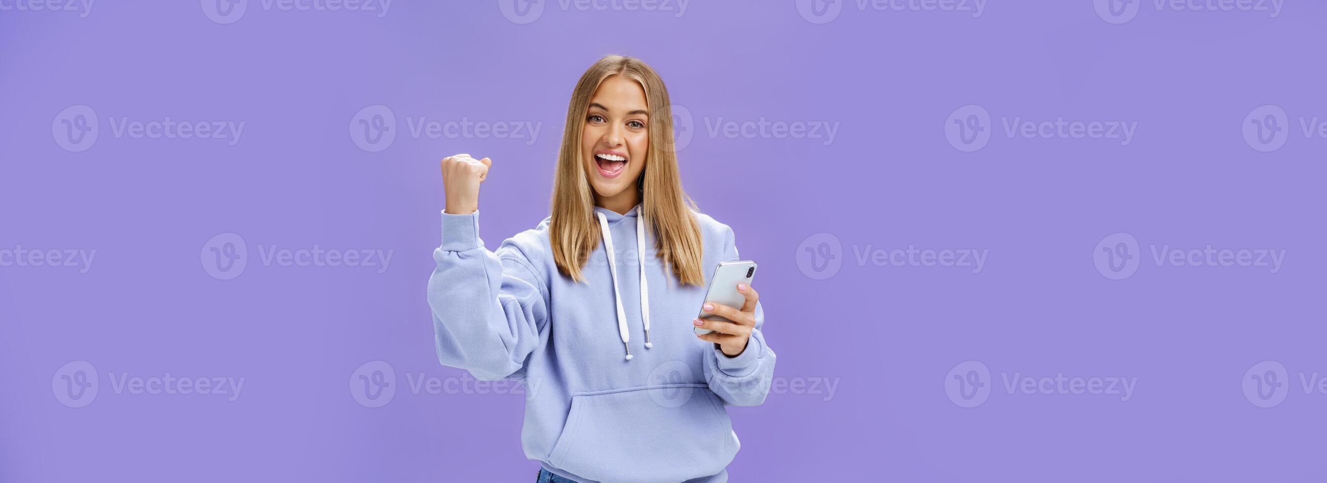 Ja wij deed het. verheugd opgewonden vrouw in capuchon verhogen vuist in viering en succes gebaar glimlachen breed zegevieren Holding smartphone reageren naar positief nieuws in mobiele telefoon over- Purper muur foto