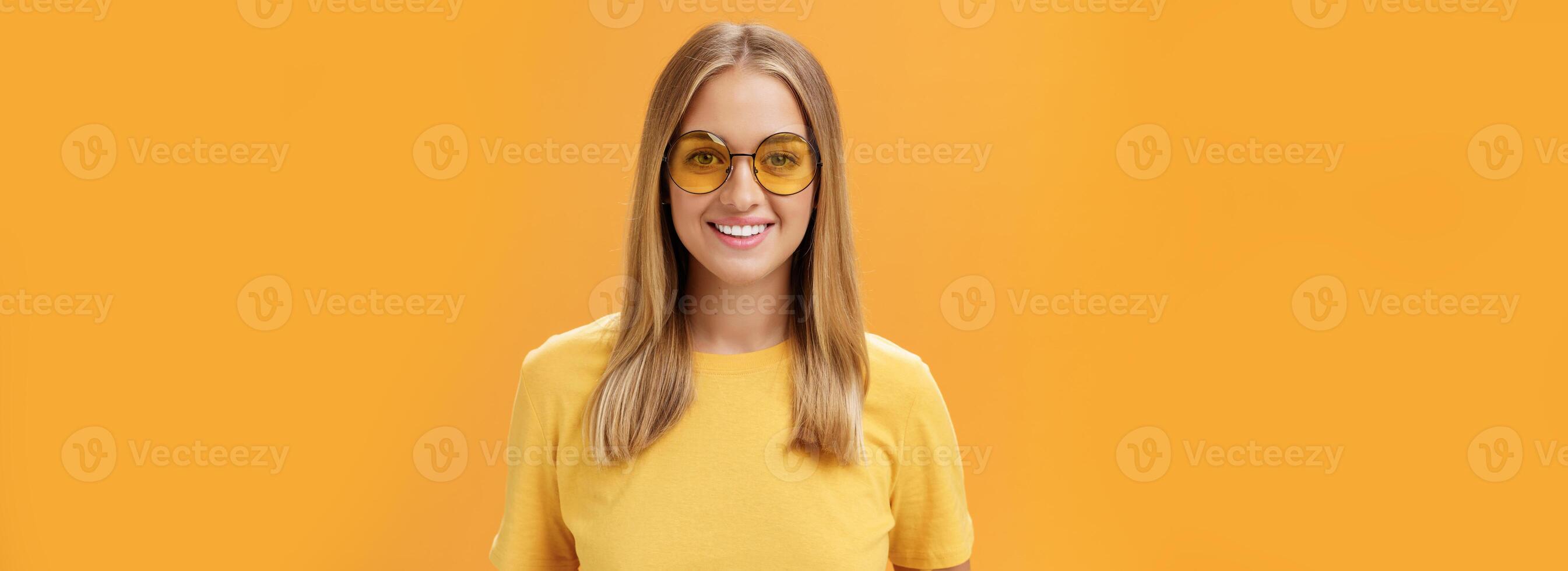elegant zelfverzekerd charmant Europese vrouw met eerlijk haar- en gebruind huid in geel t-shirt en zonnebril glimlachen breed geamuseerd en vrolijk staren Bij camera poseren tegen oranje achtergrond foto