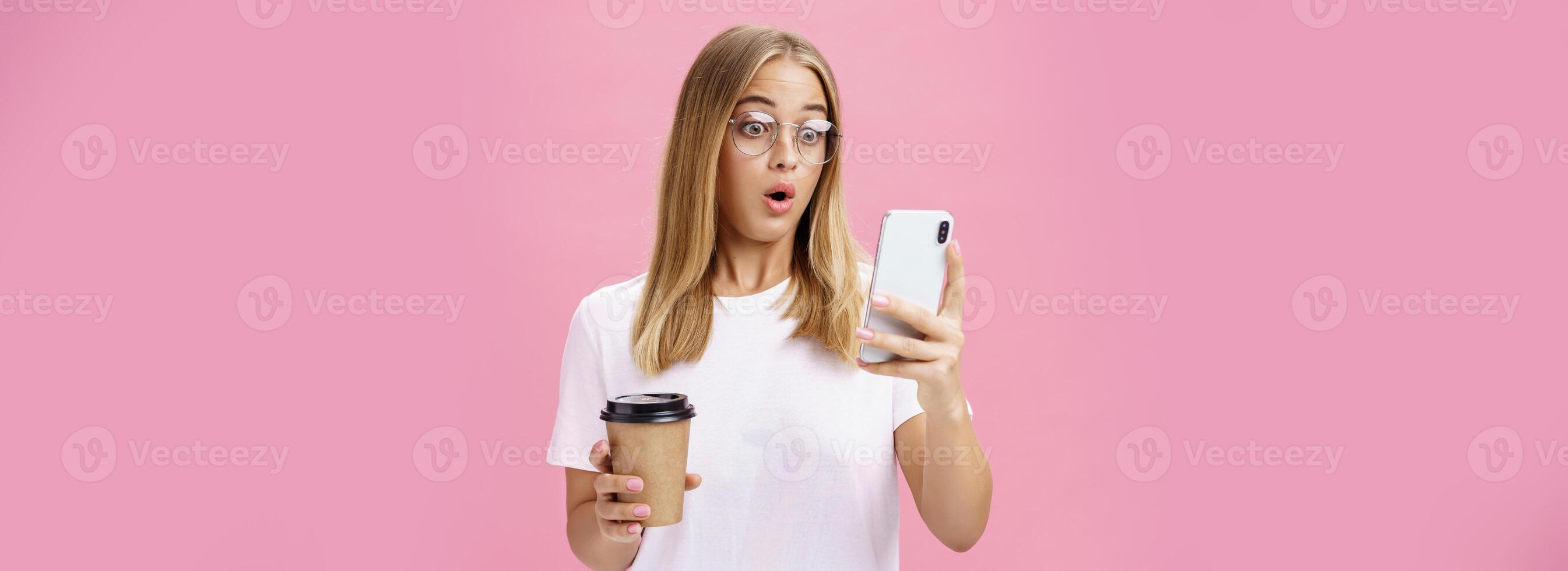 vrouw drinken koffie wezen geschokt door ontvangen bericht reageren Aan verbijsterend nieuws vouwen lippen hijgen op zoek verbaasd en onder de indruk Bij smartphone scherm Holding papier beker, poseren over- roze muur foto