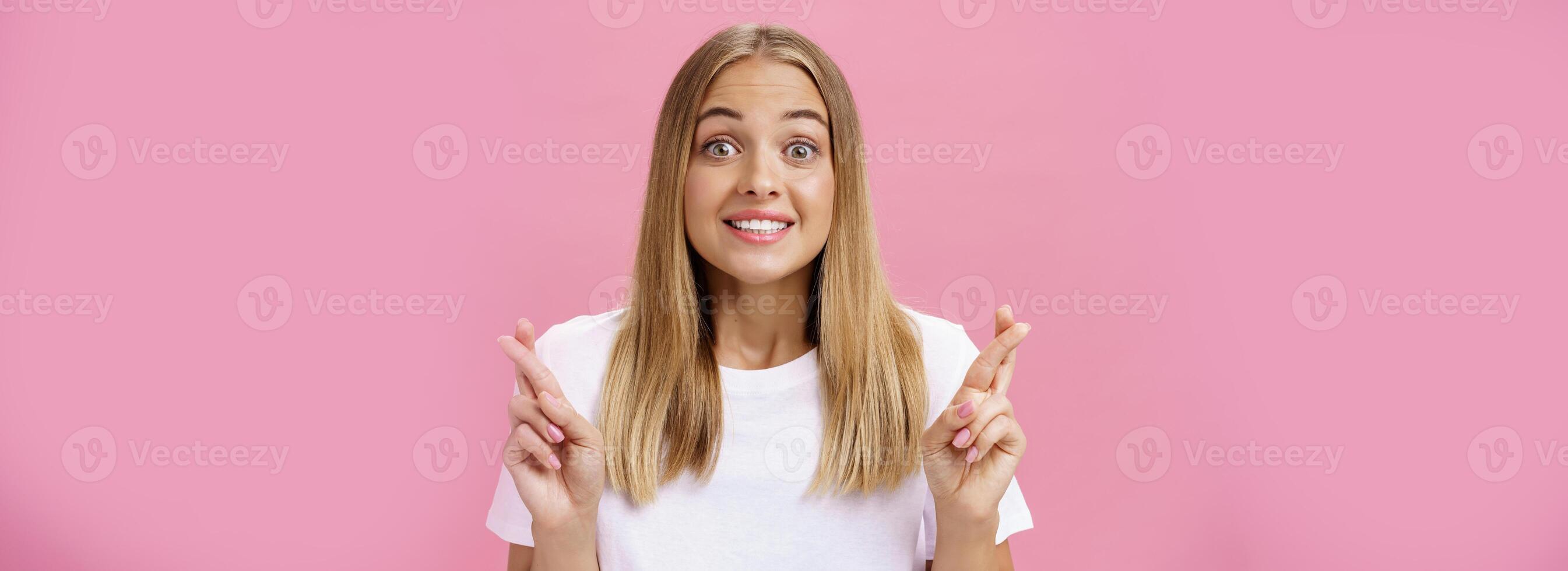 optimistisch hoopvol bekrachtigd meisje met gebruind huid en Rechtdoor eerlijk haar- looien opgewonden en gelukkig Bij camera glimlachen met hoop kruispunt vingers voor mooi zo geluk maken wens poseren tegen roze muur foto