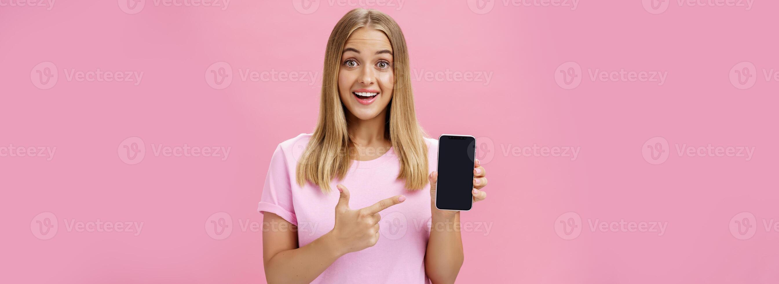 vrolijk aantrekkelijk en aangenaam vrouw bevorderen koel app of smartphone Holding mobiele telefoon en richten Bij apparaat scherm glimlachen geamuseerd en onder de indruk staand over- roze achtergrond foto