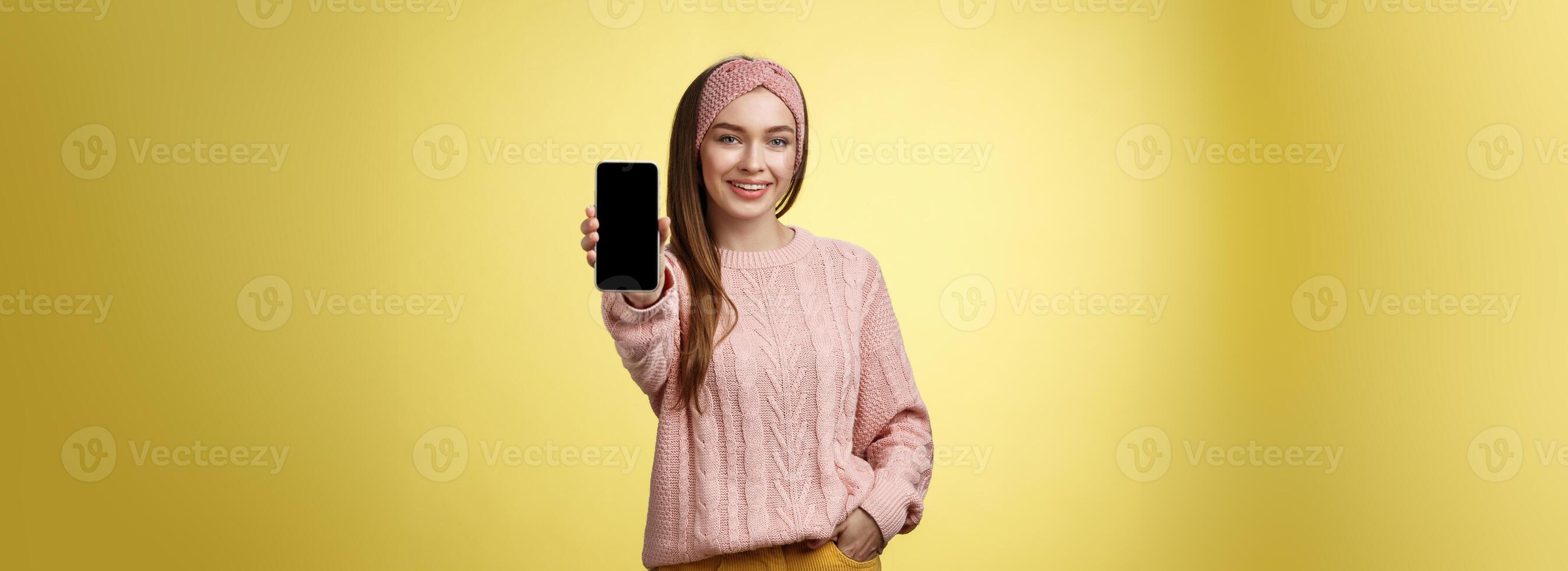 het beste smartphone hier u Gaan. charmant uitgaand jong mooi vrouw in gebreid trui, hoofdband uitbreiden hand- met telefoon tonen apparaatje scherm glimlachen aanbevelen mobiele telefoon over- geel muur foto