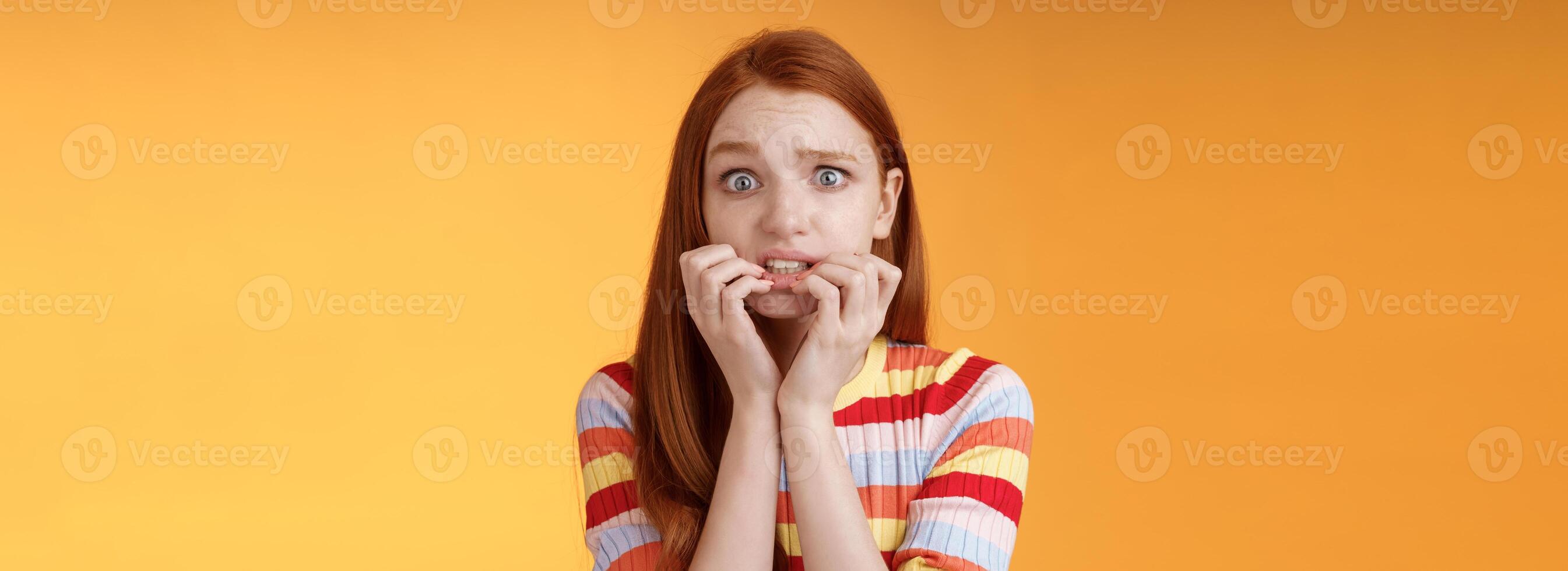 angstig onzelfverzekerd timide onzeker roodharige schattig meisje zich zorgen maken beven angst gevolgen bijten vingernagels fronsen grimassen bang, staand nerveus oranje achtergrond in afwachting eng moment foto