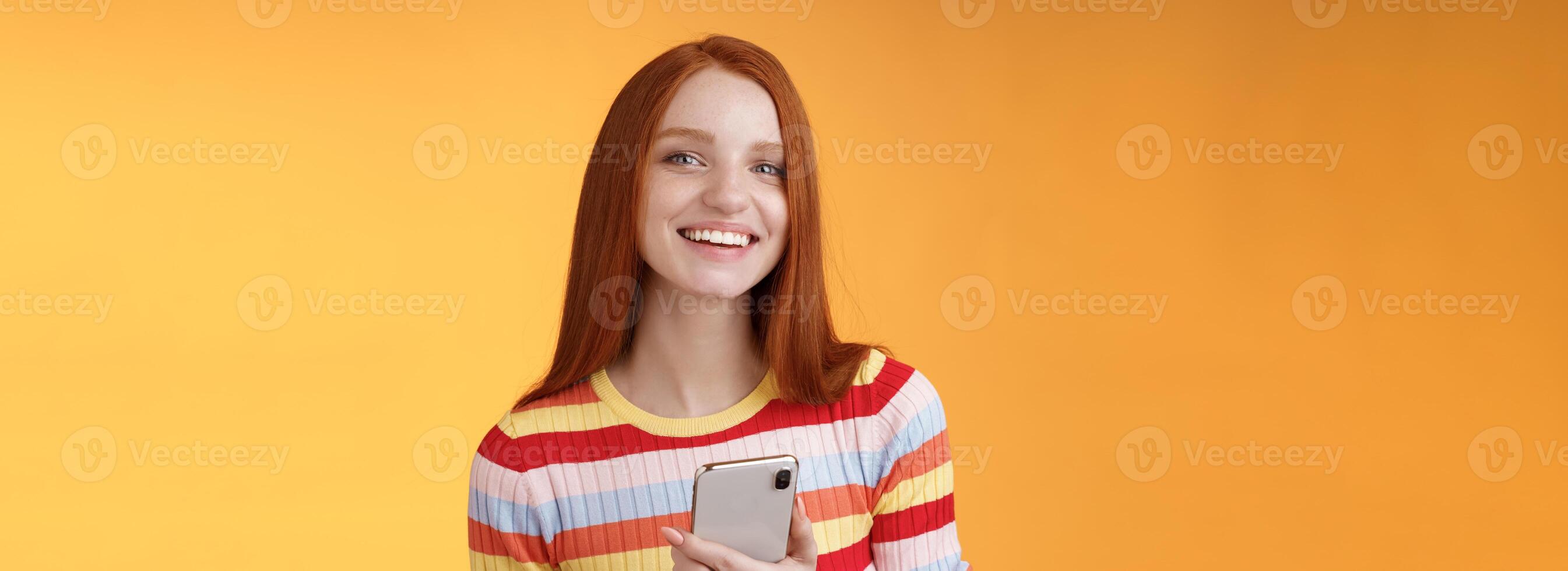 vriendelijk enthousiast jong roodharige meisje blauw ogen gebruik makend van smartphone beurt camera antwoord glimlachen breed vertellen wie verzonden bericht staand verheugd oranje achtergrond berichten, sms'en vriendje foto