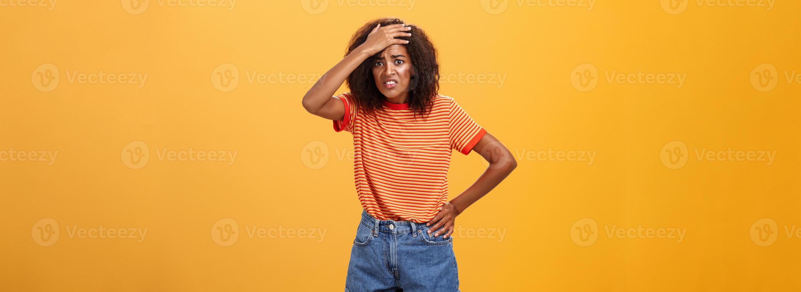 shit ik ben in probleem. bezorgd angstig schattig met een donkere huid vrouw in modieus t-shirt en shorts balanceren tanden met schuldig kijken fronsen ponsen voorhoofd hebben probleem of probleem over- oranje muur foto