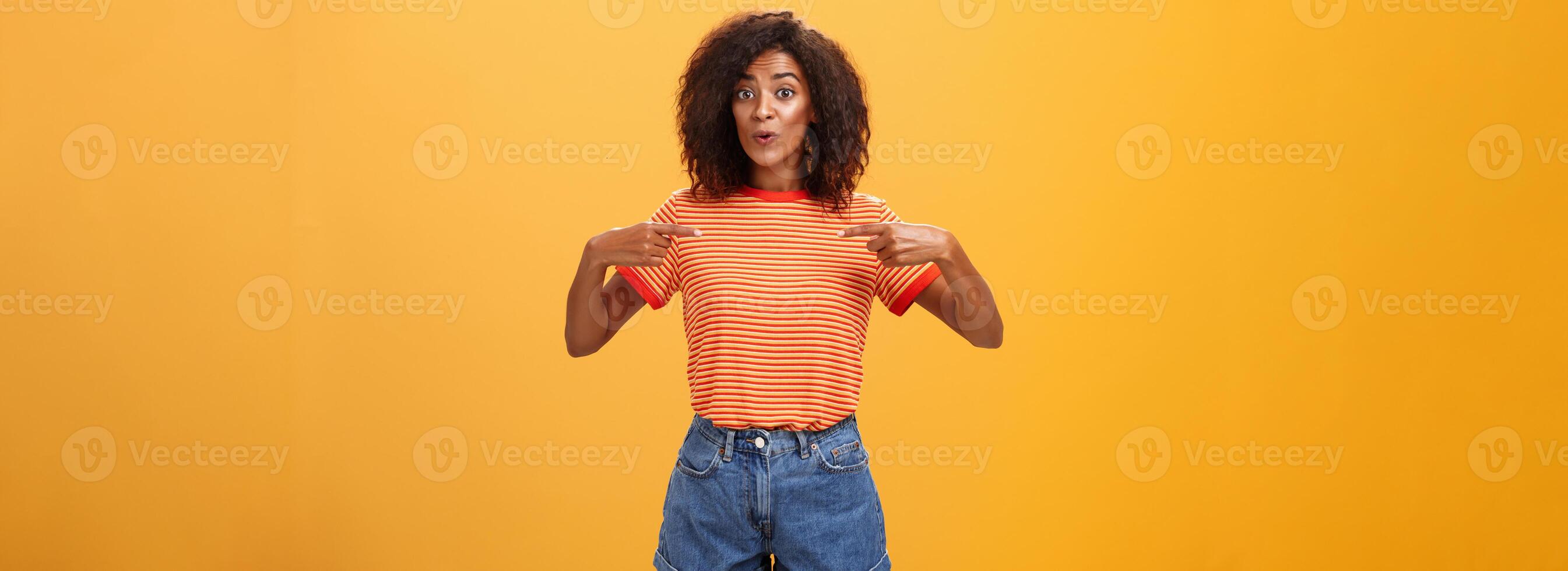 vrouw wezen verheugd vertellen vriend ze kreeg nieuw baan richten Bij haarzelf met verheugd tevreden kijken opscheppen staand gelukkig over- oranje achtergrond in elegant denim shorts en gestreept t-shirt foto