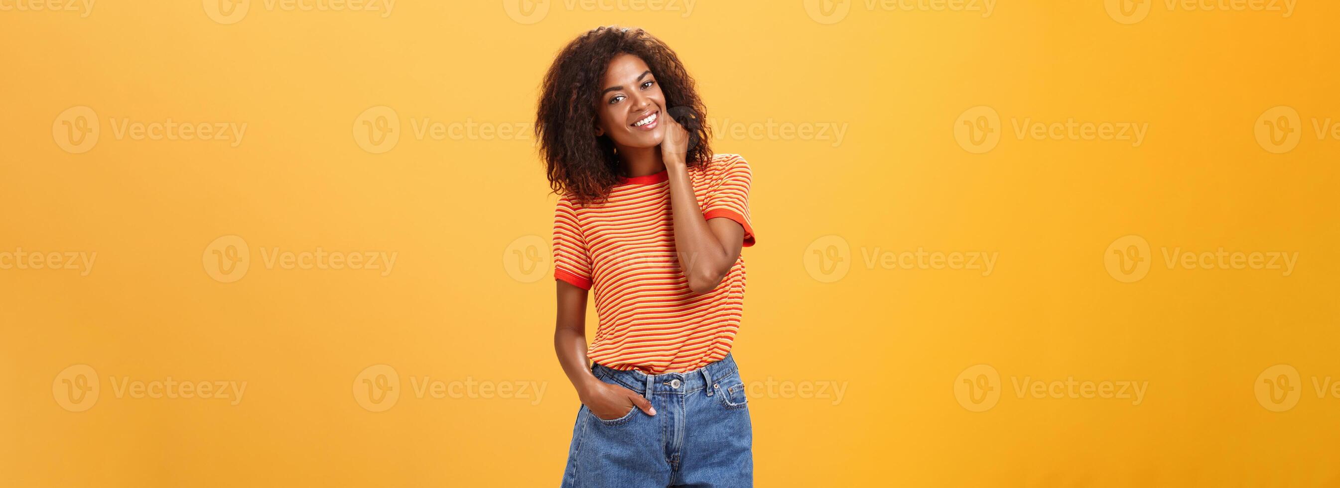 elegant Afrikaanse Amerikaans slank vrouw met gekruld medium haar- Holding hand- in denim shorts kantelen hoofd, wrijven nek terwijl glimlachen vriendelijk gevoel ongemakkelijk of verlegen in nieuw bedrijf over- oranje muur foto