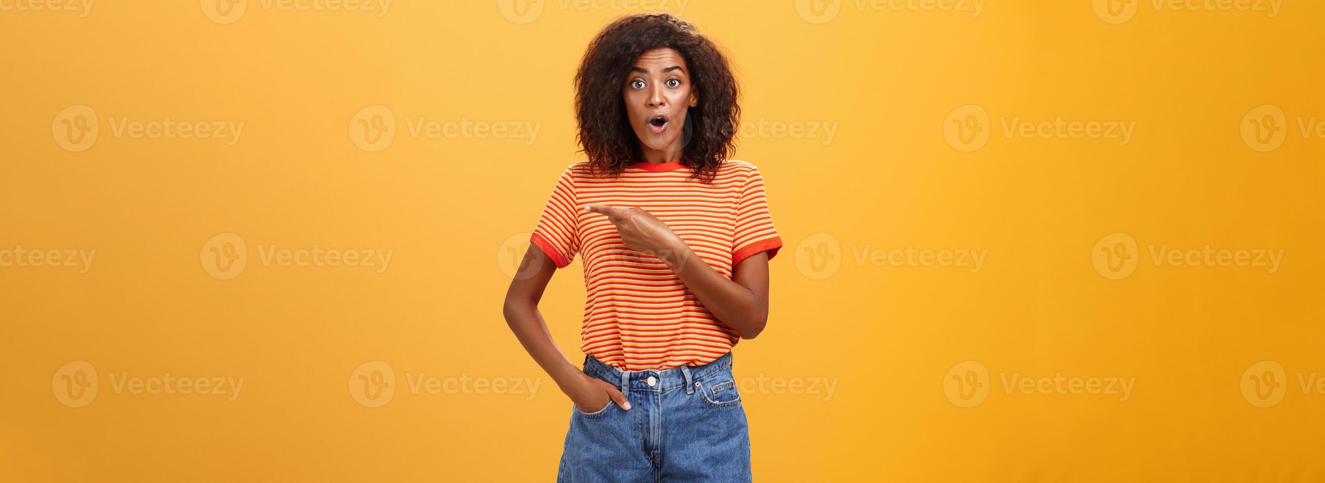 studio schot van elegant fit vrouw model- in denim shorts en t-shirt hijgen onder de indruk richten Rechtsaf terwijl vragen vraag wezen geïnteresseerd gedurende observeren masterclass over- oranje achtergrond foto