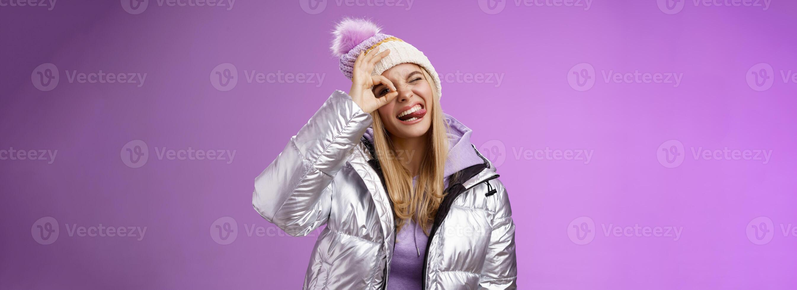 vriendelijk blij Kaukasisch blond meisje in hoed warm elegant glimmend zilver jasje kantelen hoofd gelukkig tonen tong glimlachen breed genieten van geweldig ski toevlucht vakantie op reis winter, Purper achtergrond foto