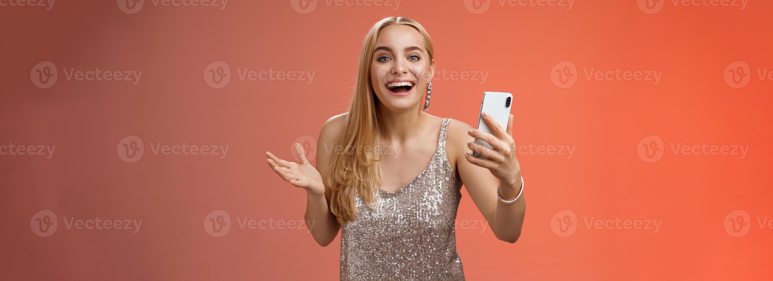 verbaasd gelukkig charmant blond meisje in zilver glinsterende elegant jurk Holding smartphone versteld staan leuk vinden geweldig resultaat Bewerk foto app glimlachen vroeg me af geamuseerd, staand rood achtergrond