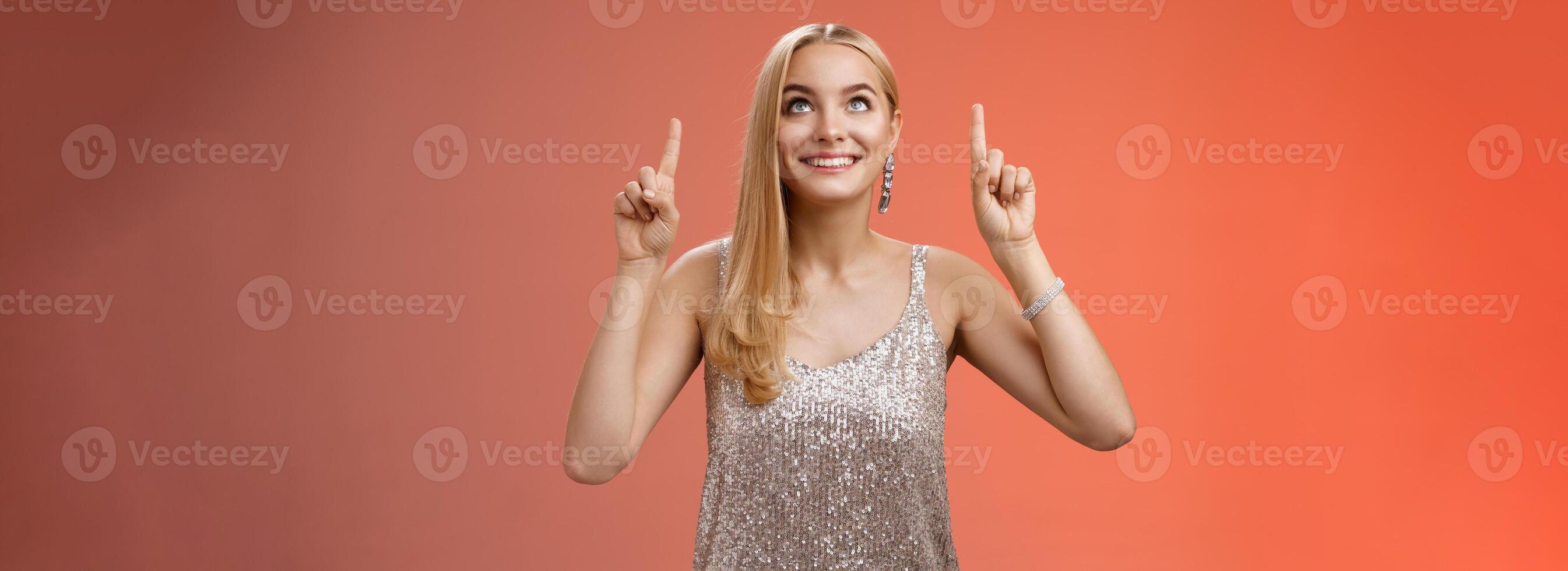 geamuseerd prachtig inschrijving elegant Europese vrouw blond lang kapsel in zilver glinsterende avond jurk kijken richten omhoog nieuwsgierig blik opwinding zien gewenst ding, staand rood achtergrond blij foto