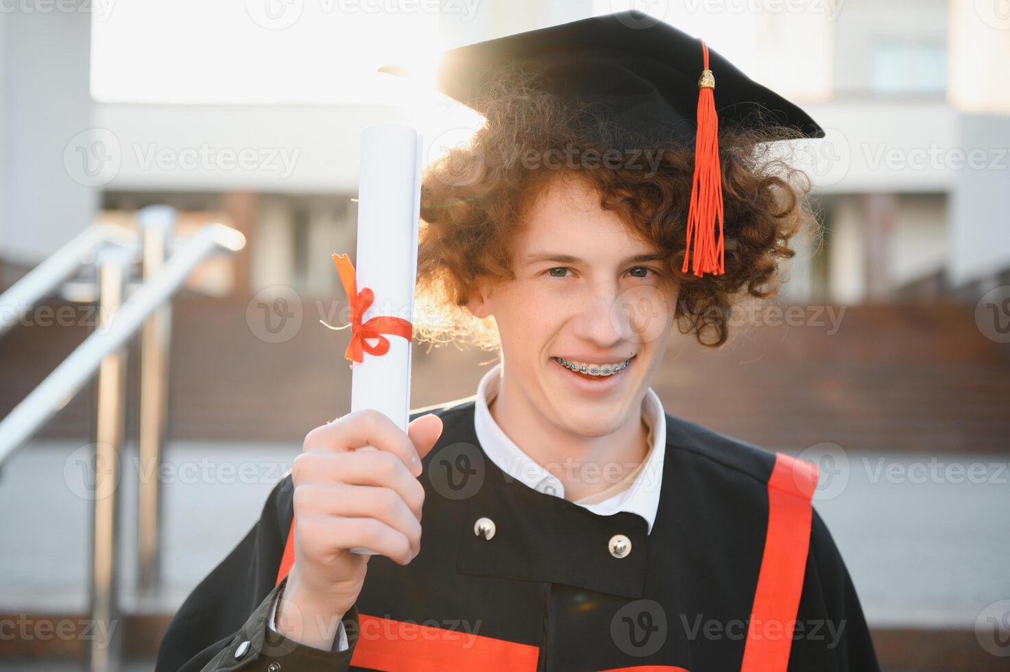 diploma uitreiking van Universiteit. jong glimlachen jongen Universiteit afstuderen in traditioneel botten en mantel staand en Holding diploma in hand- over- Universiteit gebouw achtergrond foto