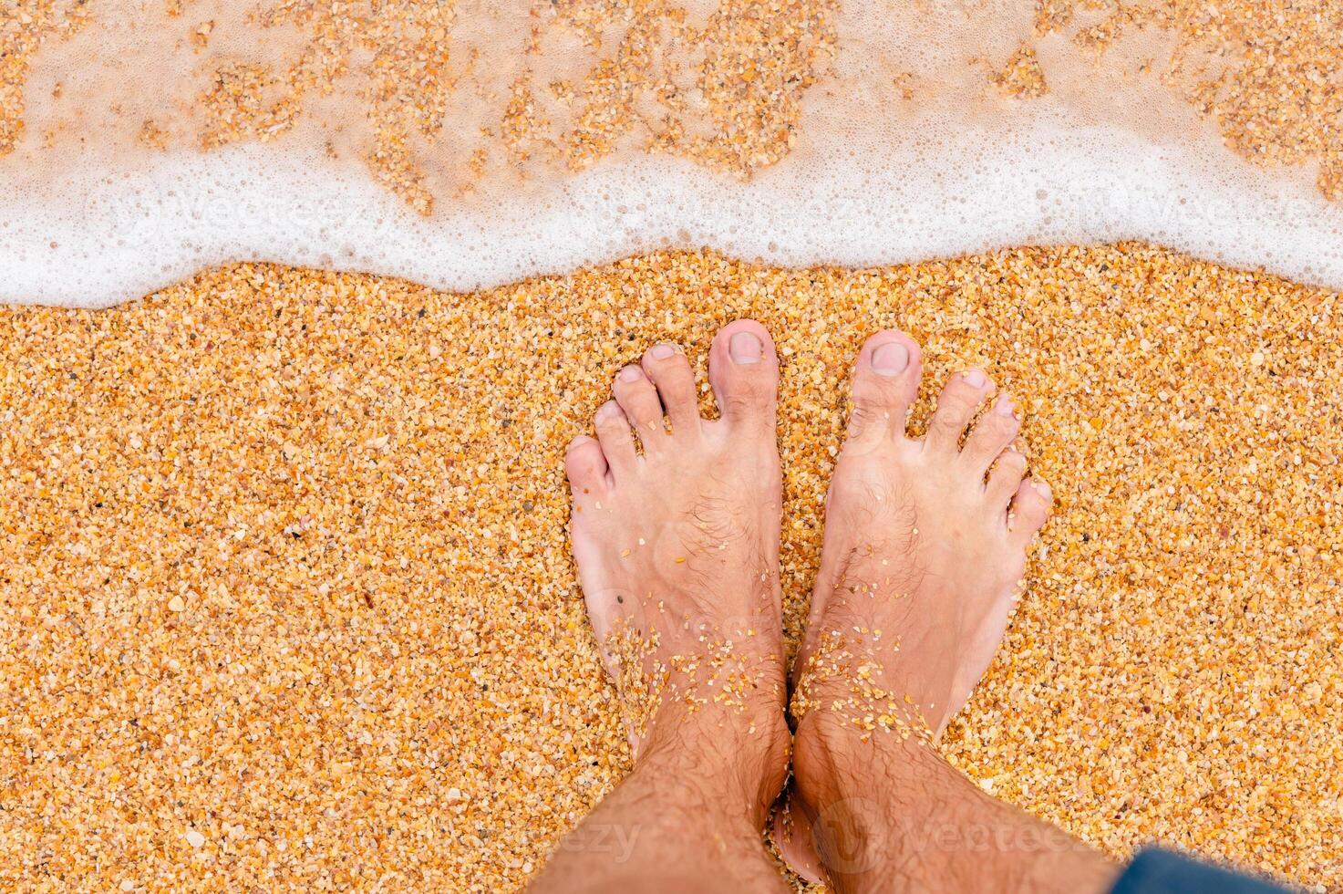 top visie van mannetje poten in blauw shorts Aan gouden zand in zonlicht. op zoek naar beneden Bij een Mens in shorts staand Aan de zee zanderig strand foto