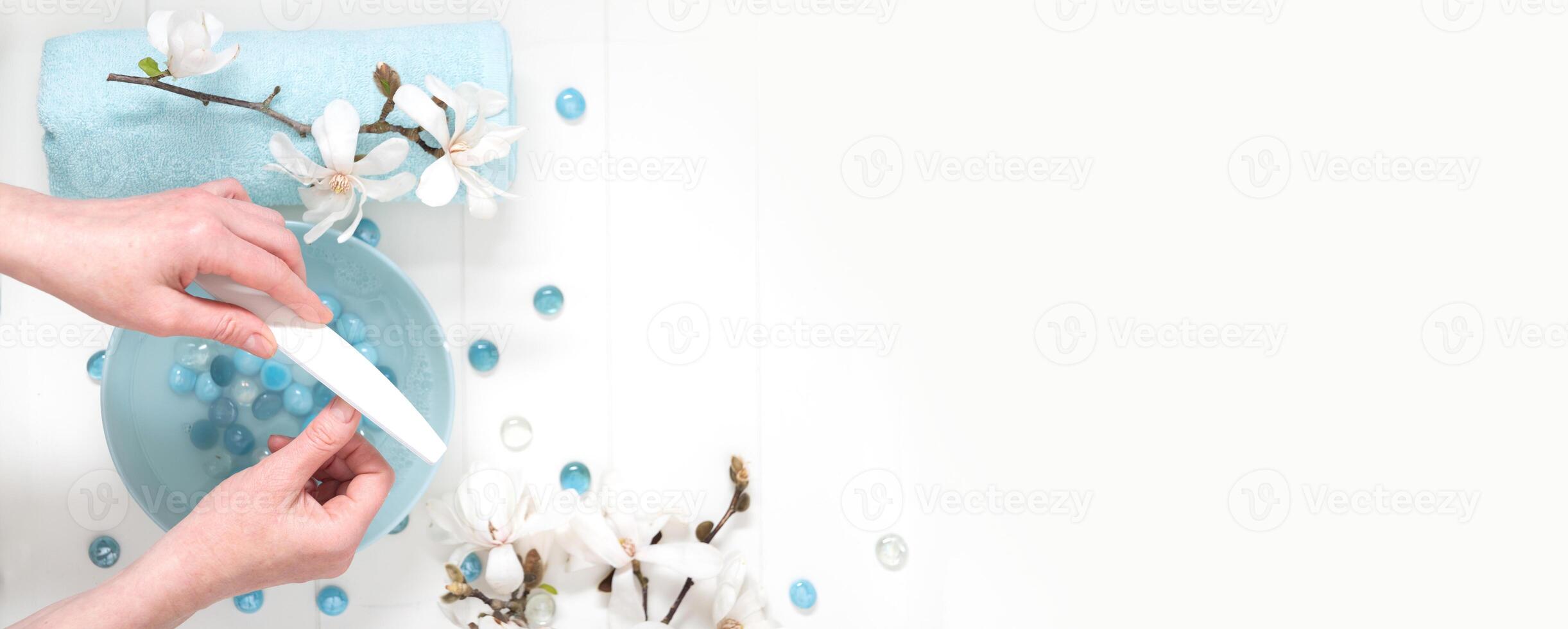 jong vrouw verscherpt haar nagels met een het dossier in de salon met een kop van voorjaar water, blauw spa stenen en wit magnolia foto