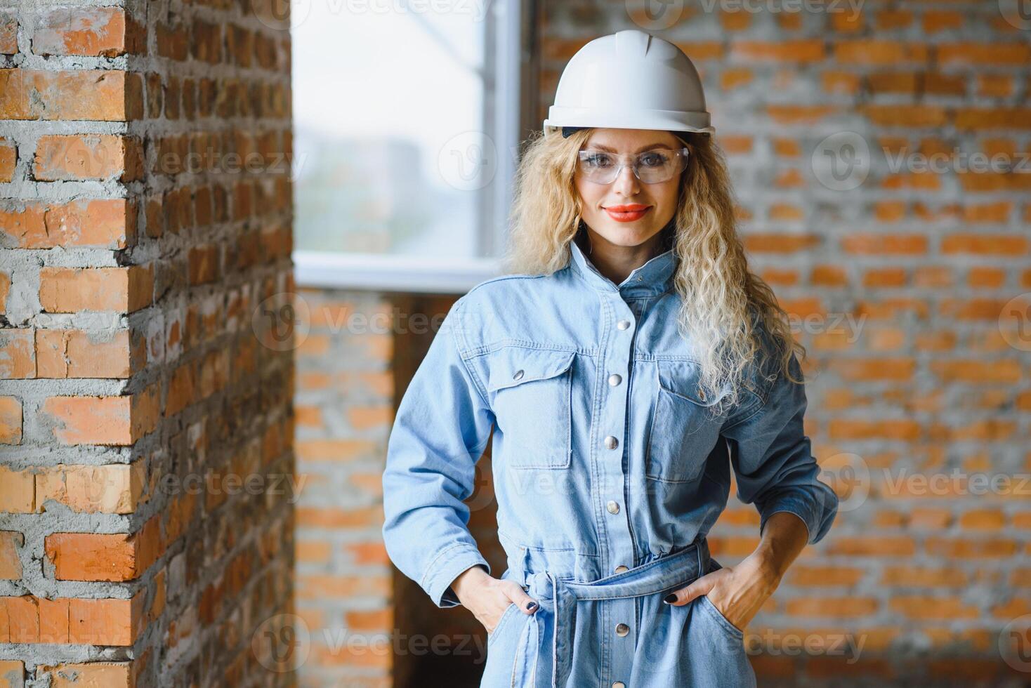 aantrekkelijk vrouw bouw arbeider in harde hoed. zelfverzekerd jong specialist in geruit blauw overhemd in jeans staand in leeg kamer. interieur ontwerp en vernieuwing onderhoud. foto