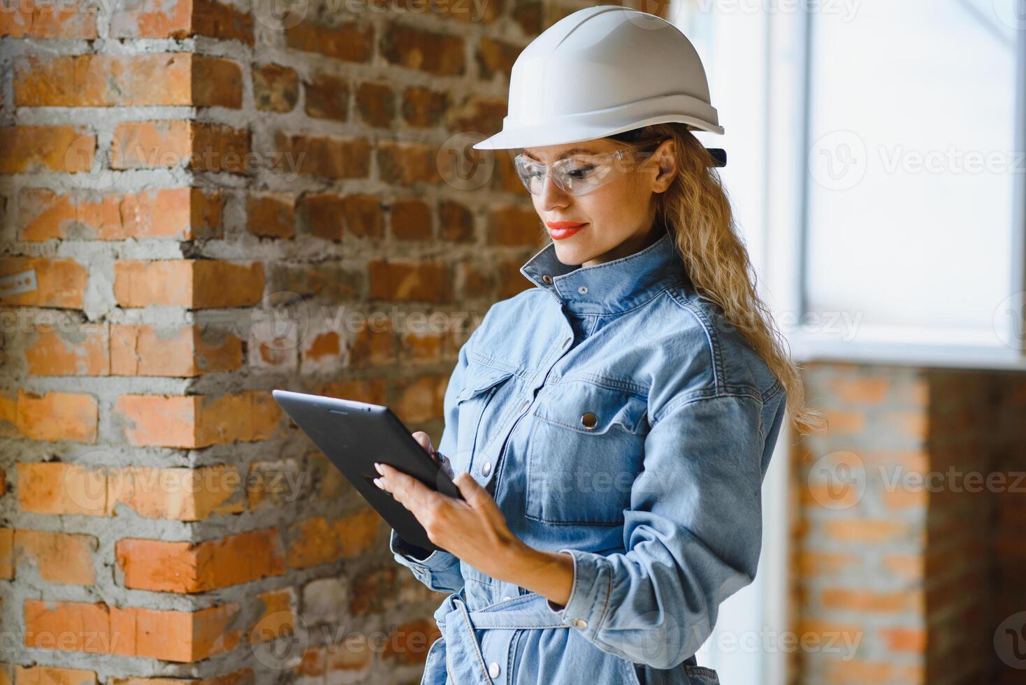 aantrekkelijk vrouw bouw arbeider in harde hoed. zelfverzekerd jong specialist in geruit blauw overhemd in jeans staand in leeg kamer. interieur ontwerp en vernieuwing onderhoud foto