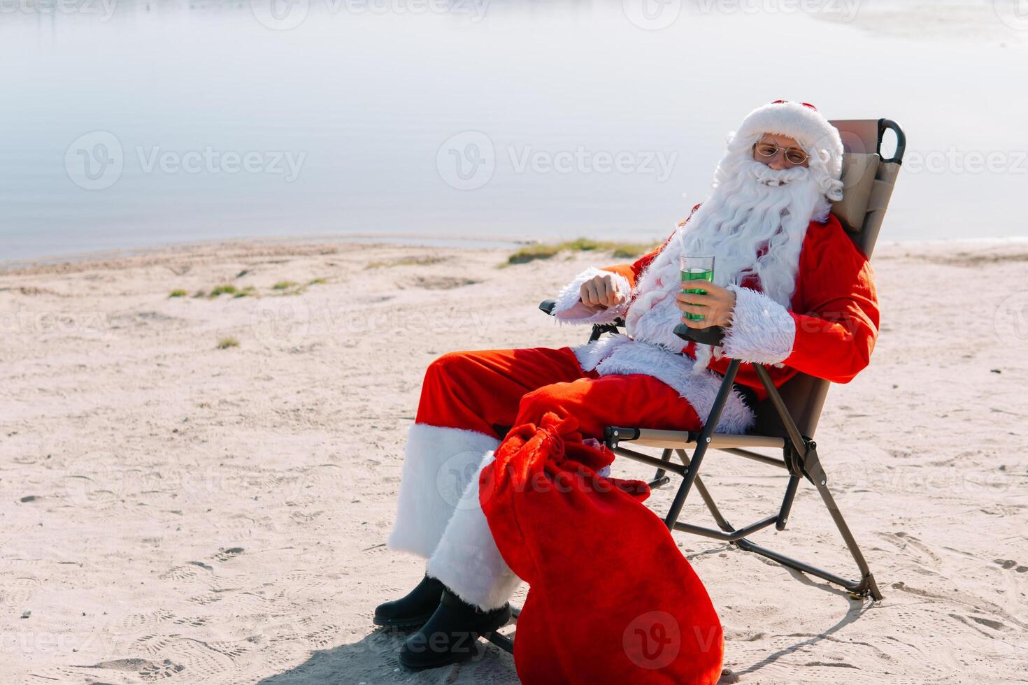 de kerstman claus in bril drankjes een cocktail terwijl aan het liegen Aan een zon ligstoel Aan de meer strand. de kerstman in de tropen foto