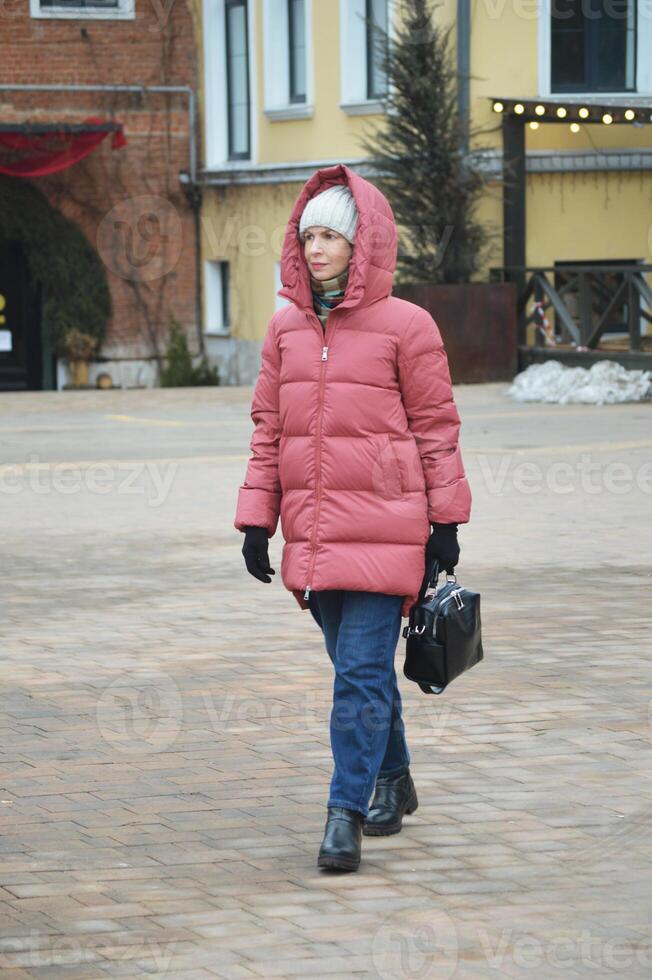 een jong elegant Kaukasisch vrouw in een naar beneden jasje, gebreid hoed en jeans wandelingen langs een stad straat in winter. foto