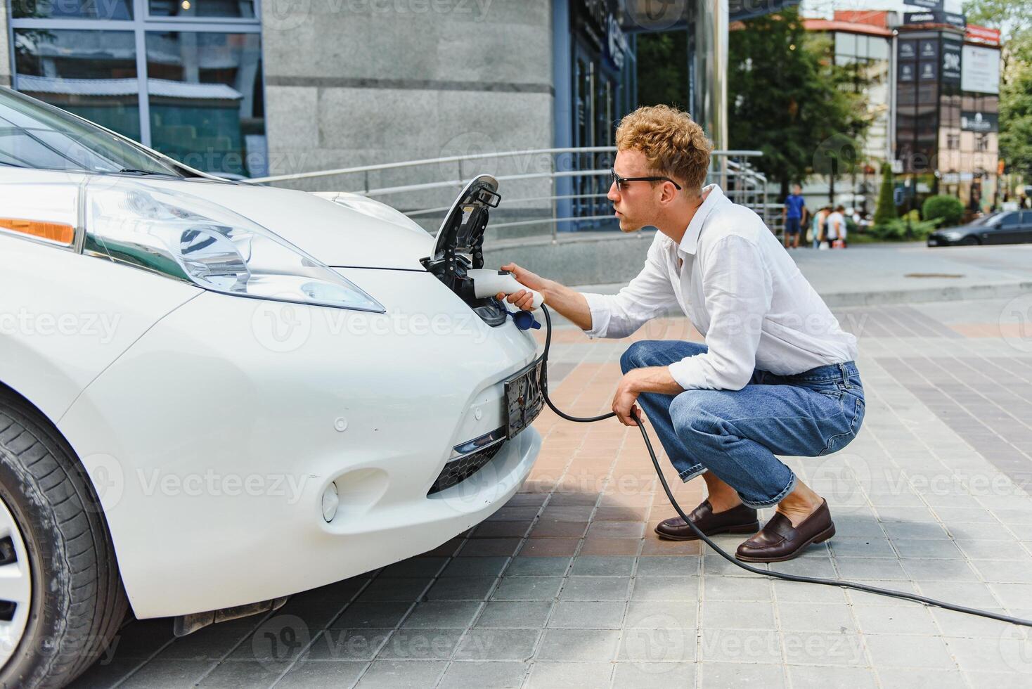 jong knap Mens Holding opladen kabel Bij elektrisch opladen station punt staand in de buurt zijn nieuw auto , op zoek tevreden. foto