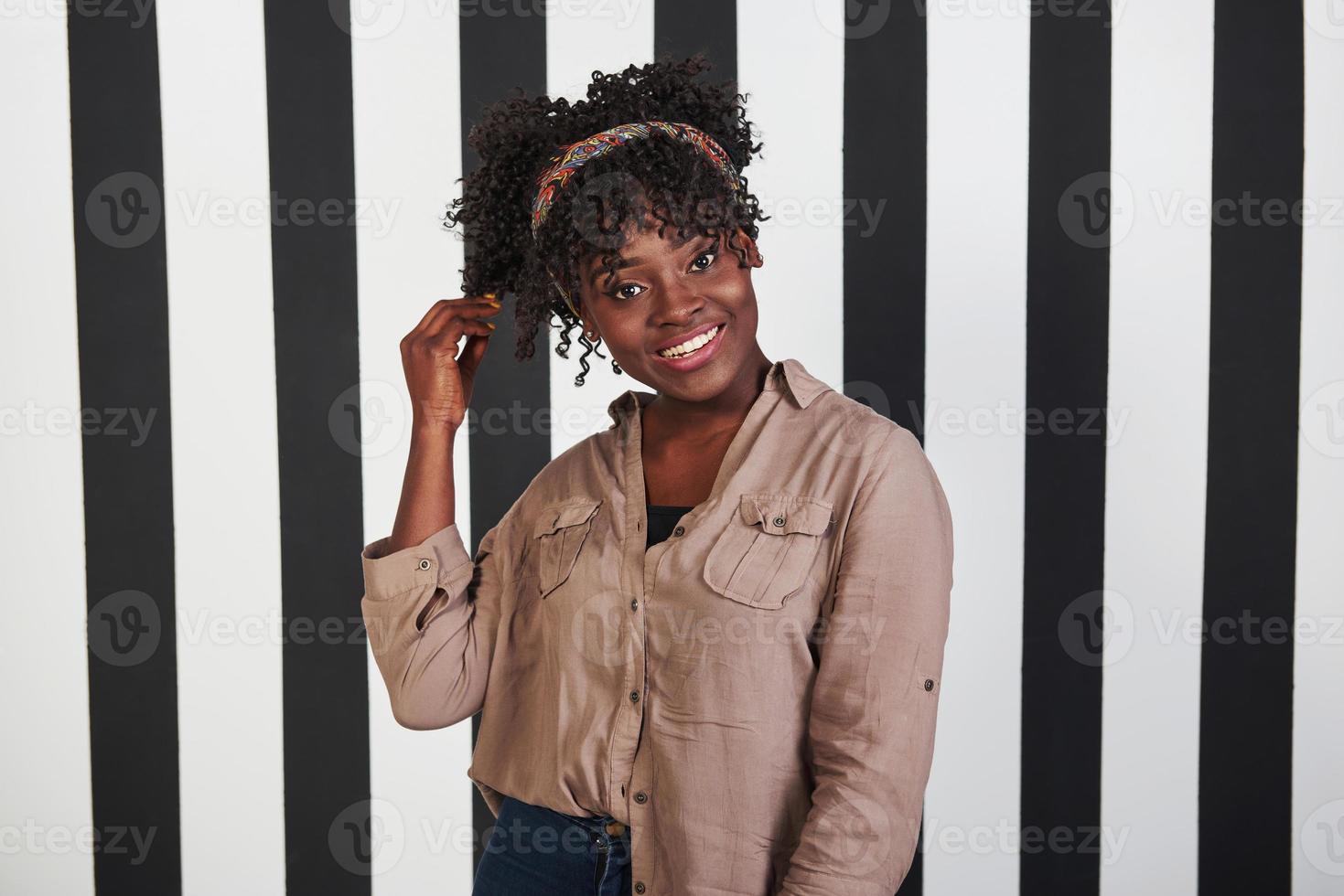 geglimlacht afro-amerikaans meisje staat en raakt haar haar aan in de studio met verticale witte en zwarte lijnen op de achtergrond foto