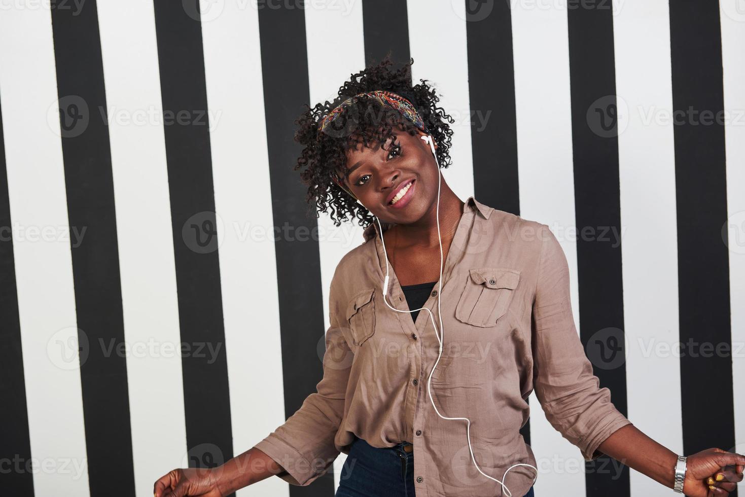 vrouw luistert muziek in een koptelefoon en danst erop. geglimlacht afro-amerikaans meisje staat in de studio met verticale witte en zwarte lijnen op de achtergrond foto