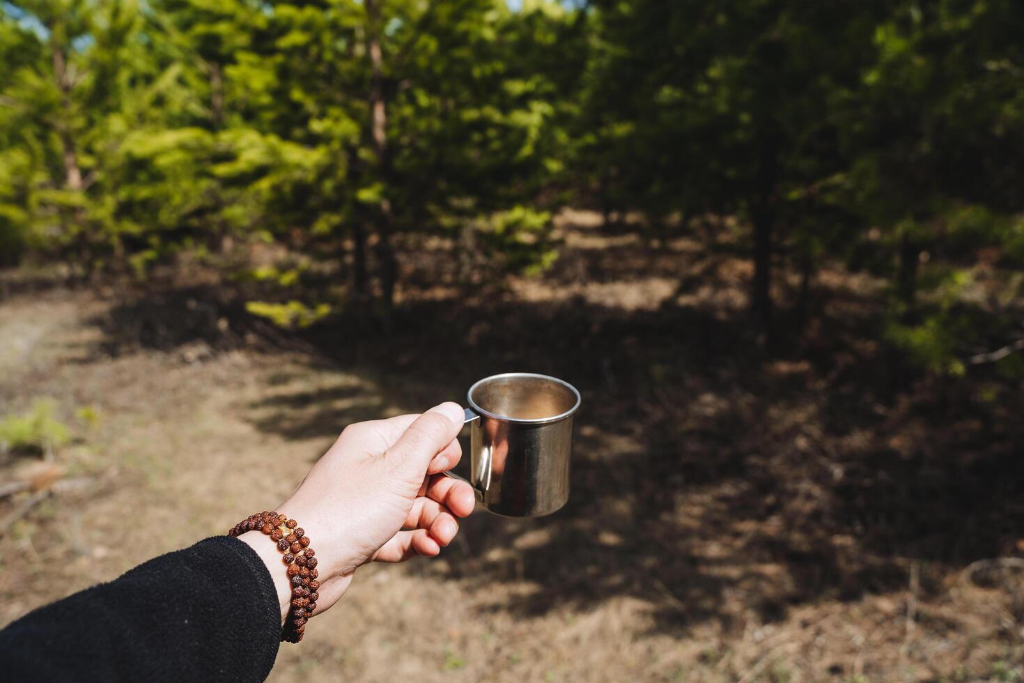 een man's hand- houdt een mok van thee tegen de achtergrond van de Woud, een metaal mok voor koffie naar drinken Aan een wandeltocht, gerechten in de Woud, natuur pijnboom zon, rudraksha armband. foto