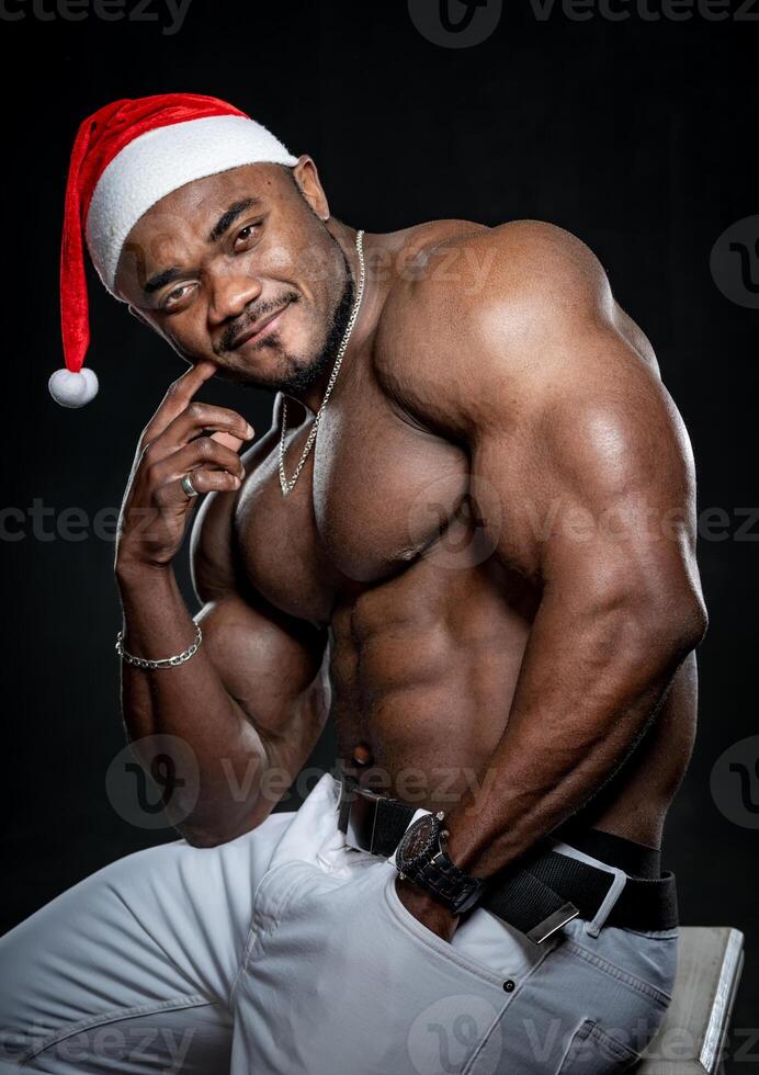 taille omhoog portret van de Afrikaanse Amerikaans bodybuilder Mens met zonder shirt gespierd torso. mannetje vervelend Kerstmis hoed geïsoleerd Aan zwart achtergrond foto