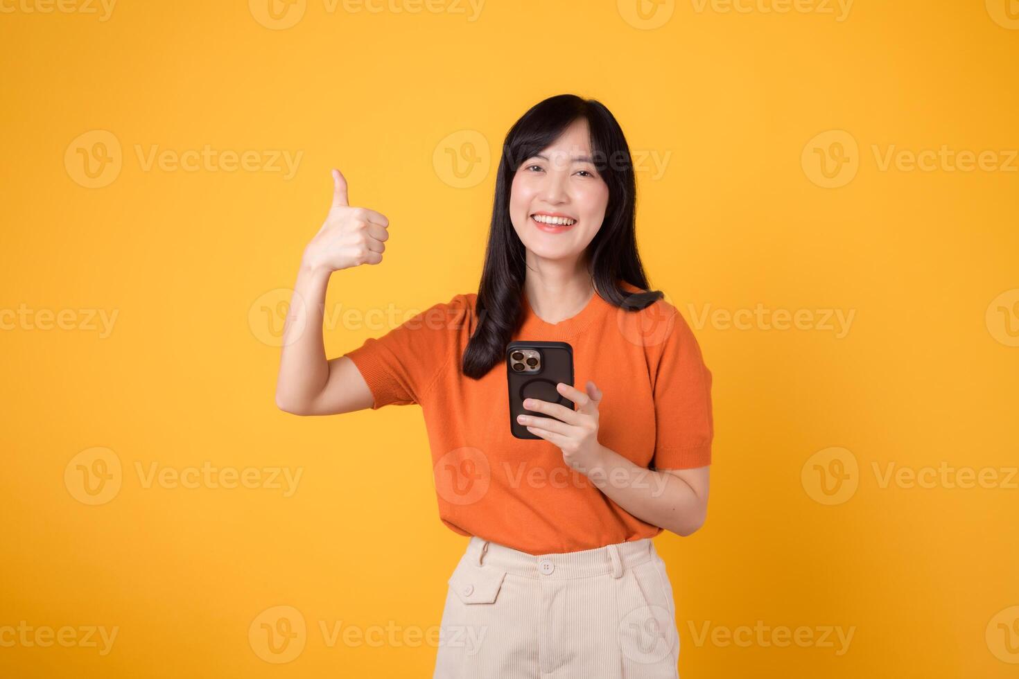 energiek jong Aziatisch vrouw in haar jaren '30, vervelend oranje shirt, gebruik makend van smartphone met duim omhoog hand- teken Aan levendig geel achtergrond. nieuw mobiel app concept. foto