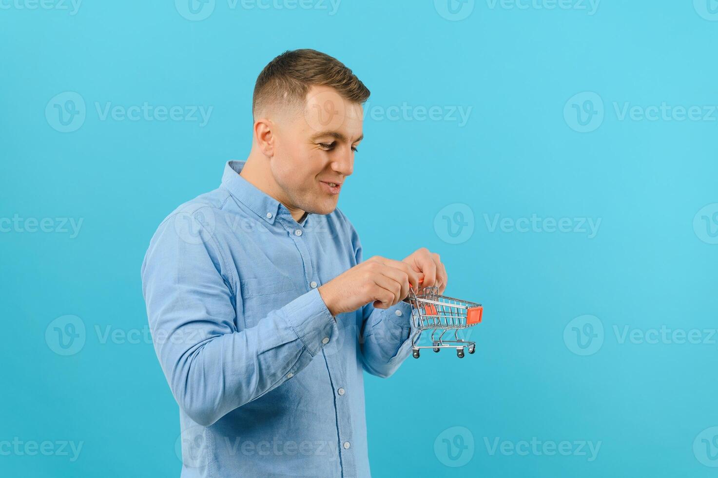 jong glimlachen Mens Holding een mini boodschappen doen kar Aan een blauw achtergrond. verkoop concept. plaats voor tekst. foto