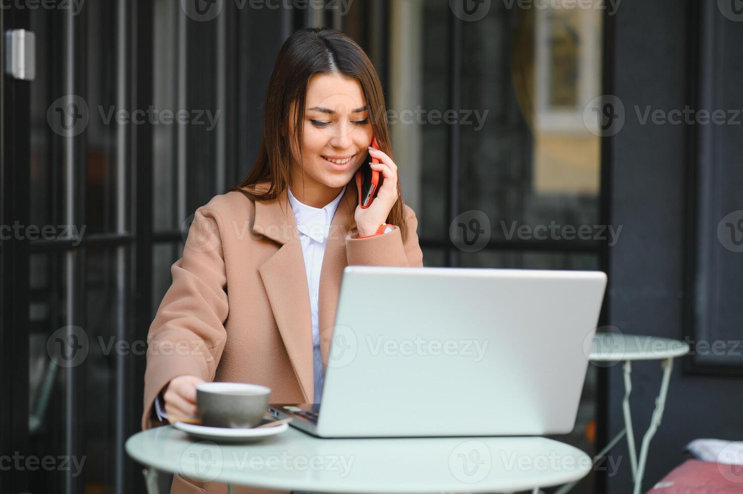 jong gelukkig bedrijf dame looks Bij smartphone scherm met toothy glimlach terwijl zittend buiten Aan terras met laptop en koffie, werken van een afstand online controle rapporten en krijgen klaar voor vergadering foto