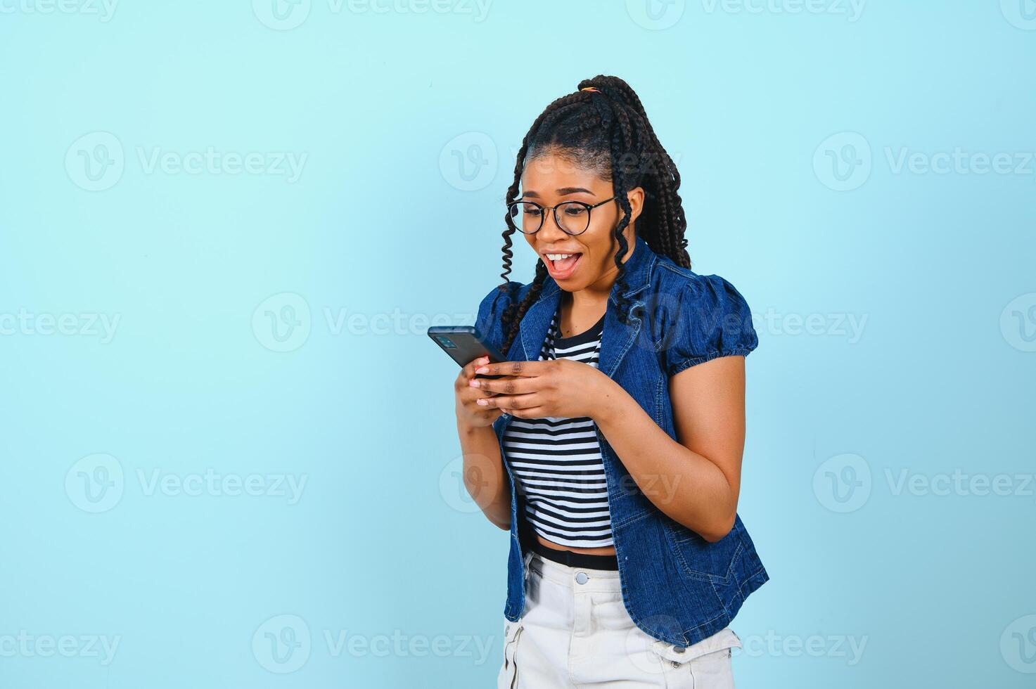 gelukkig vrolijk afro Amerikaans vrouw looks Bij scherm van slim telefoon geniet online chatten types tekst bericht surft sociaal netwerken gekleed terloops poses tegen blauw achtergrond. technologie concept foto