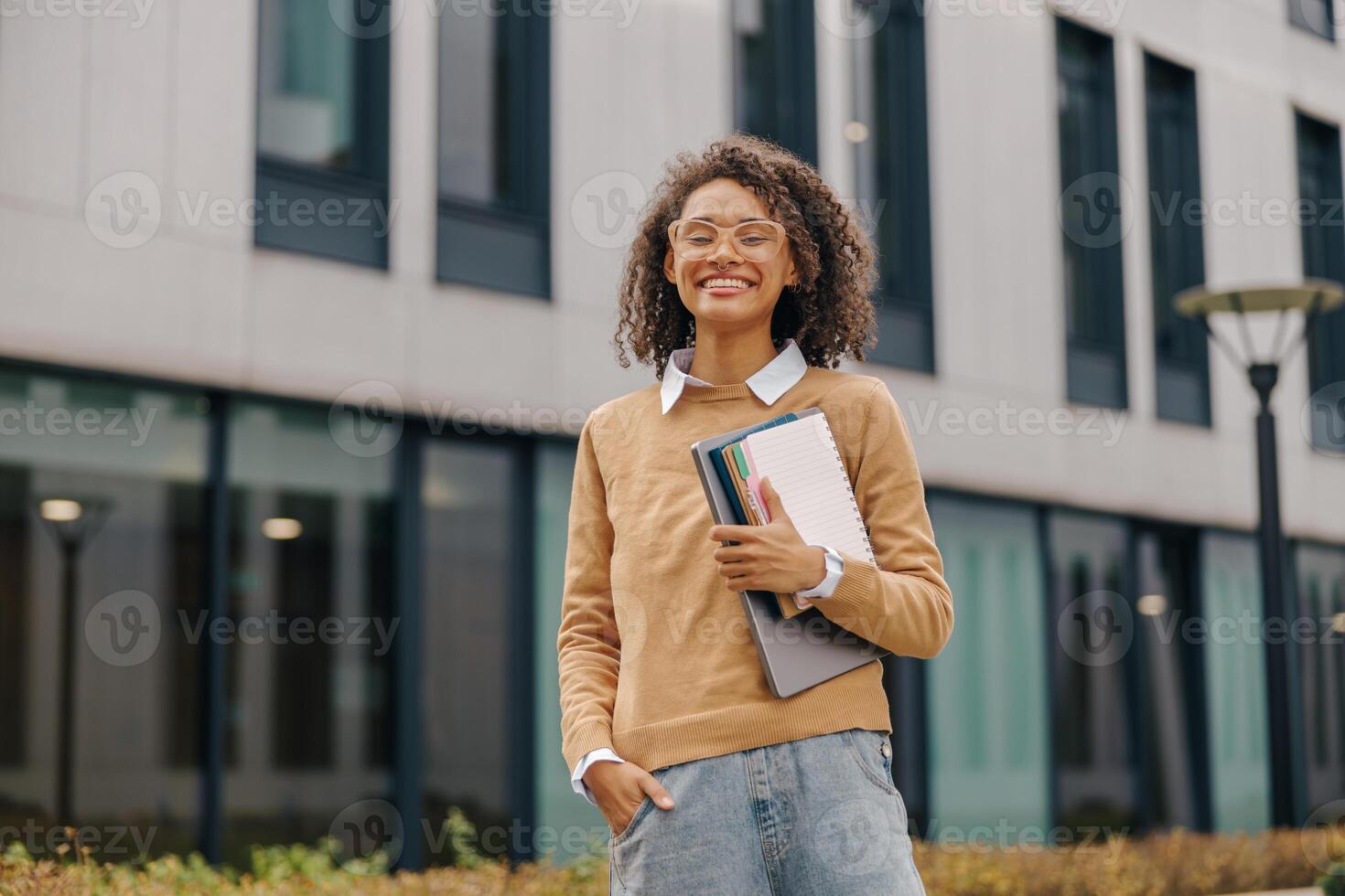 jong glimlachen vrouw leerling staand met laptop en Notitie stootkussens Aan modern gebouw achtergronden foto