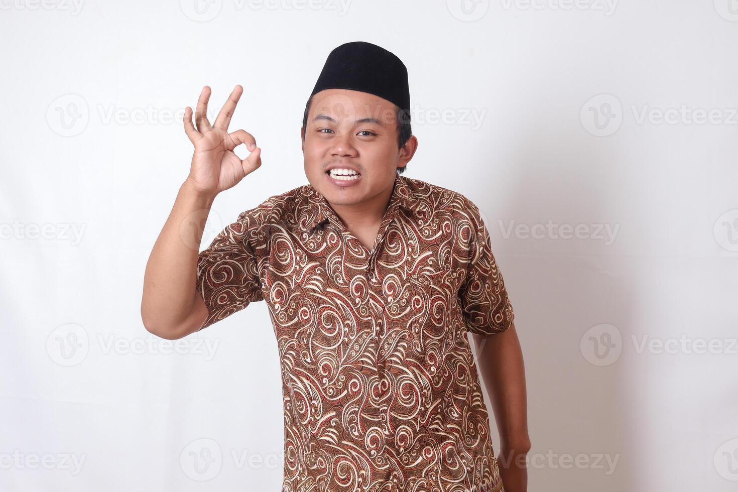 portret van opgewonden Aziatisch Mens vervelend batik overhemd en songkok tonen OK hand- gebaar en glimlachen op zoek Bij camera. geïsoleerd beeld Aan grijs achtergrond foto