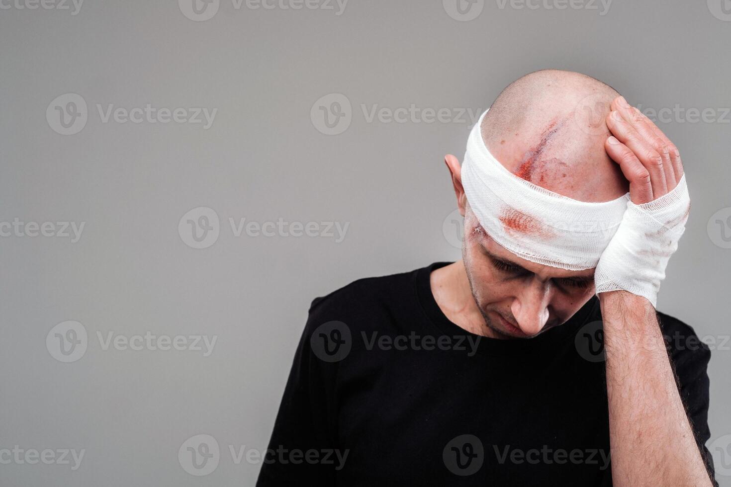 een gehavend en gehavend Mens in een zwart t-shirt staat tegen een grijs achtergrond, Holding zijn pijn doen hoofd met zijn handen verpakt in de omgeving van het foto