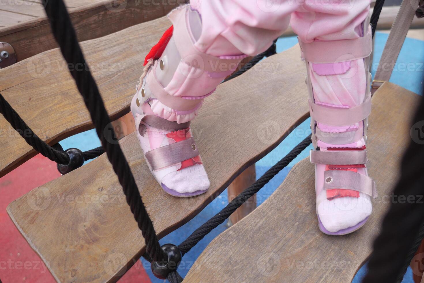 een weinig meisje in orthese schoenen kruisen een houten brug kind cerebraal verlamming onbekwaamheid. foto