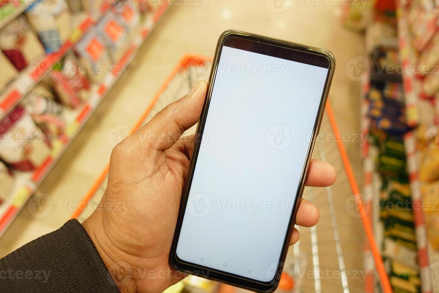 hand- Holding mobiel telefoon met wit scherm terwijl boodschappen doen Bij supermarkt. foto