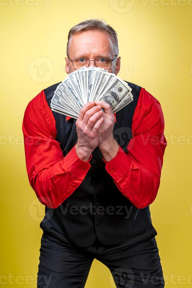 portret van gelukkig en wit tanden glimlach senior oud bedrijf Mens Holding geld in handen, gekleed in rood shirt, geïsoleerd Aan geel achtergrond. menselijk emoties en gelaats uitdrukkingen foto