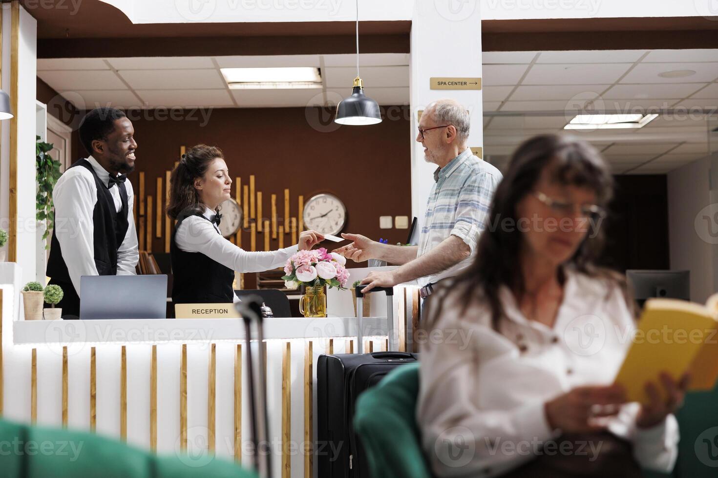 ouder Kaukasisch Mens registreert Bij hotel lobby voorkant bureau en krijgt kamer sleutel kaart van de conciërge. ouderen mannetje toerist ontvangen Super goed onderhoud van medewerkers terwijl oud vrouw zit Aan sofa met boek. foto