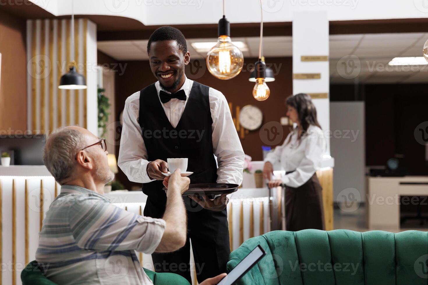 Afrikaanse Amerikaans werknemer portie koffie naar bejaarde mannetje toerist met tablet in luxe hotel ontvangst. senior Mens Holding een digitaal apparaat en ontvangen een kop van thee van toevlucht ober in lounge Oppervlakte. foto