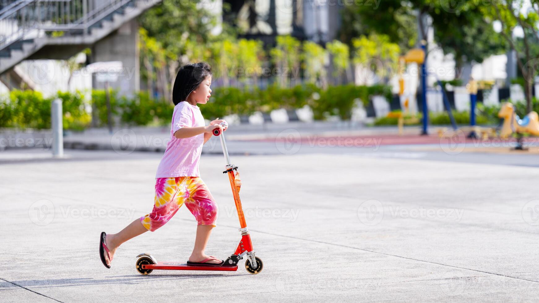 kant visie van Aziatisch kind meisje hebben pret rijden een rood scooter Bij een speelplaats, warm zonlicht, in zomer of voorjaar tijd, kopiëren ruimte, vrije tijd activiteiten, oefening kind, kinderen oud zes jaren oud. foto
