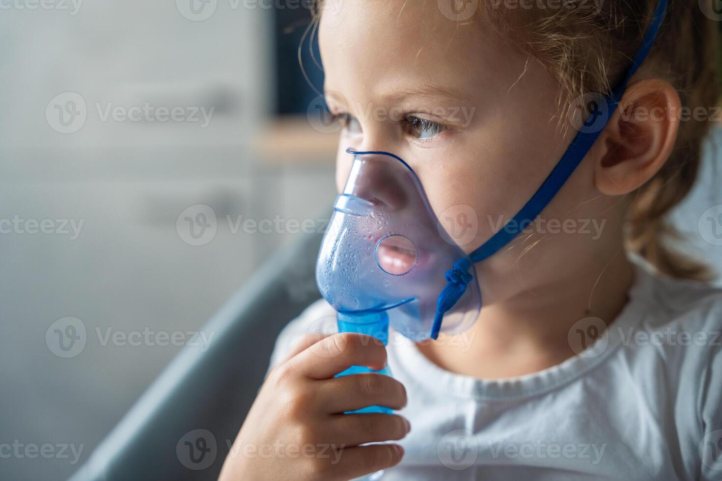 dichtbij omhoog visie van weinig meisje zijn zittend en Holding een vernevelaar masker leunend tegen de gezicht, luchtweg behandeling concept. hoog kwaliteit foto