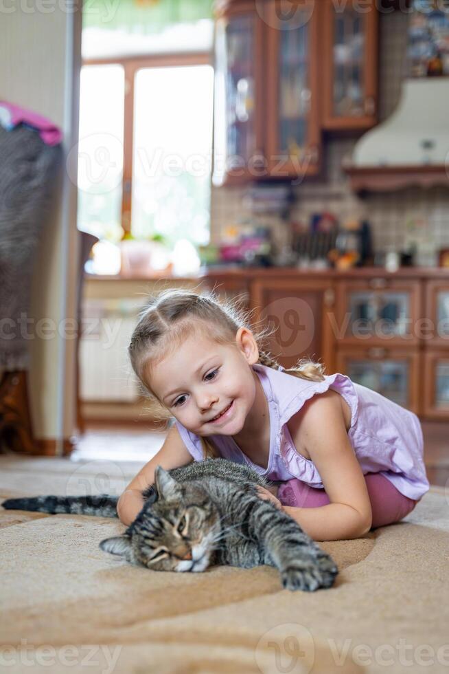 schattig weinig meisje met grijs kat aan het liegen Aan tapijt in huis leven kamer foto