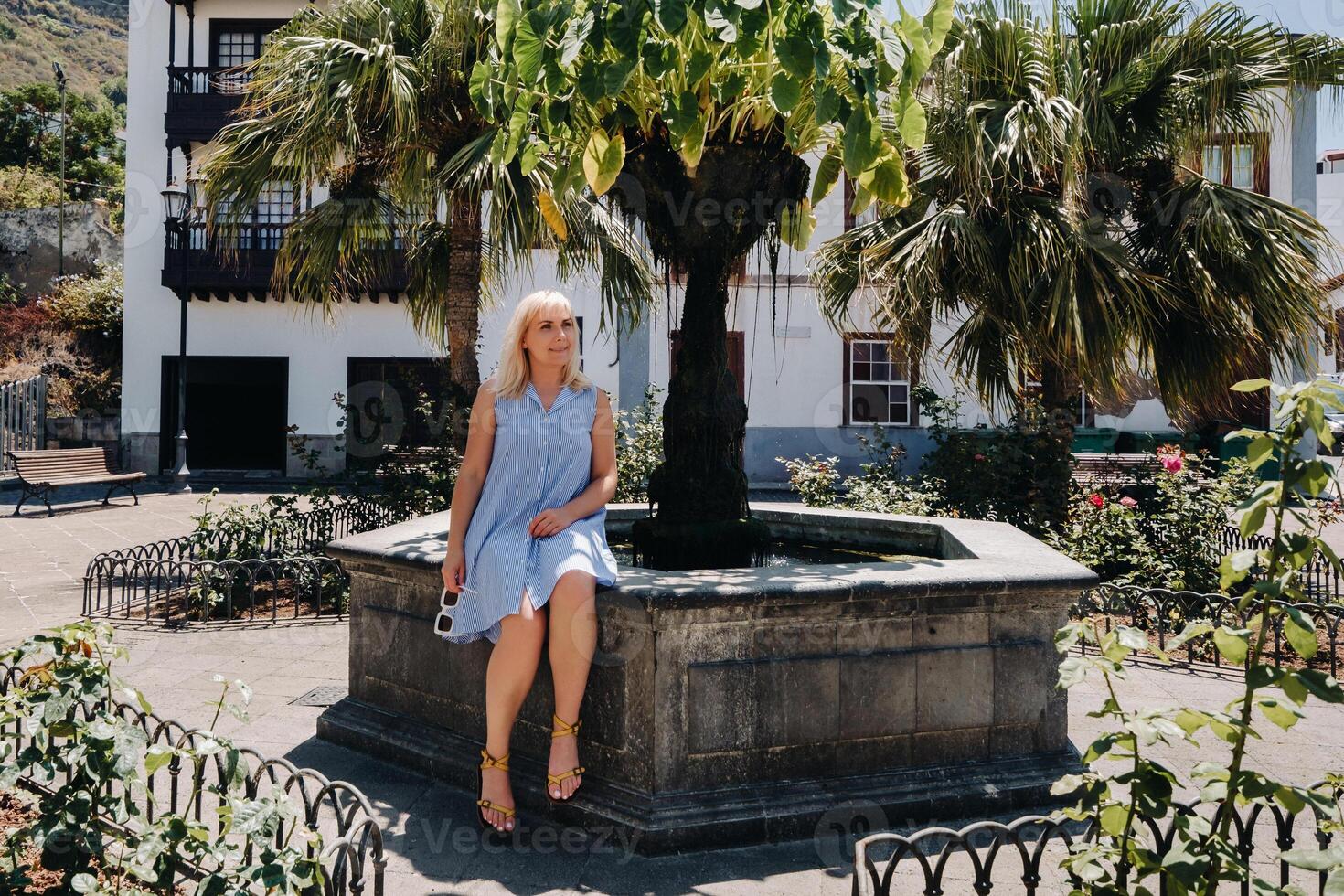 een blond vrouw in een zonnejurk zit Aan de straat van de oud stad- van icod de los Vinos Aan de eiland van tenerife.spanje, kanarie eilanden. foto